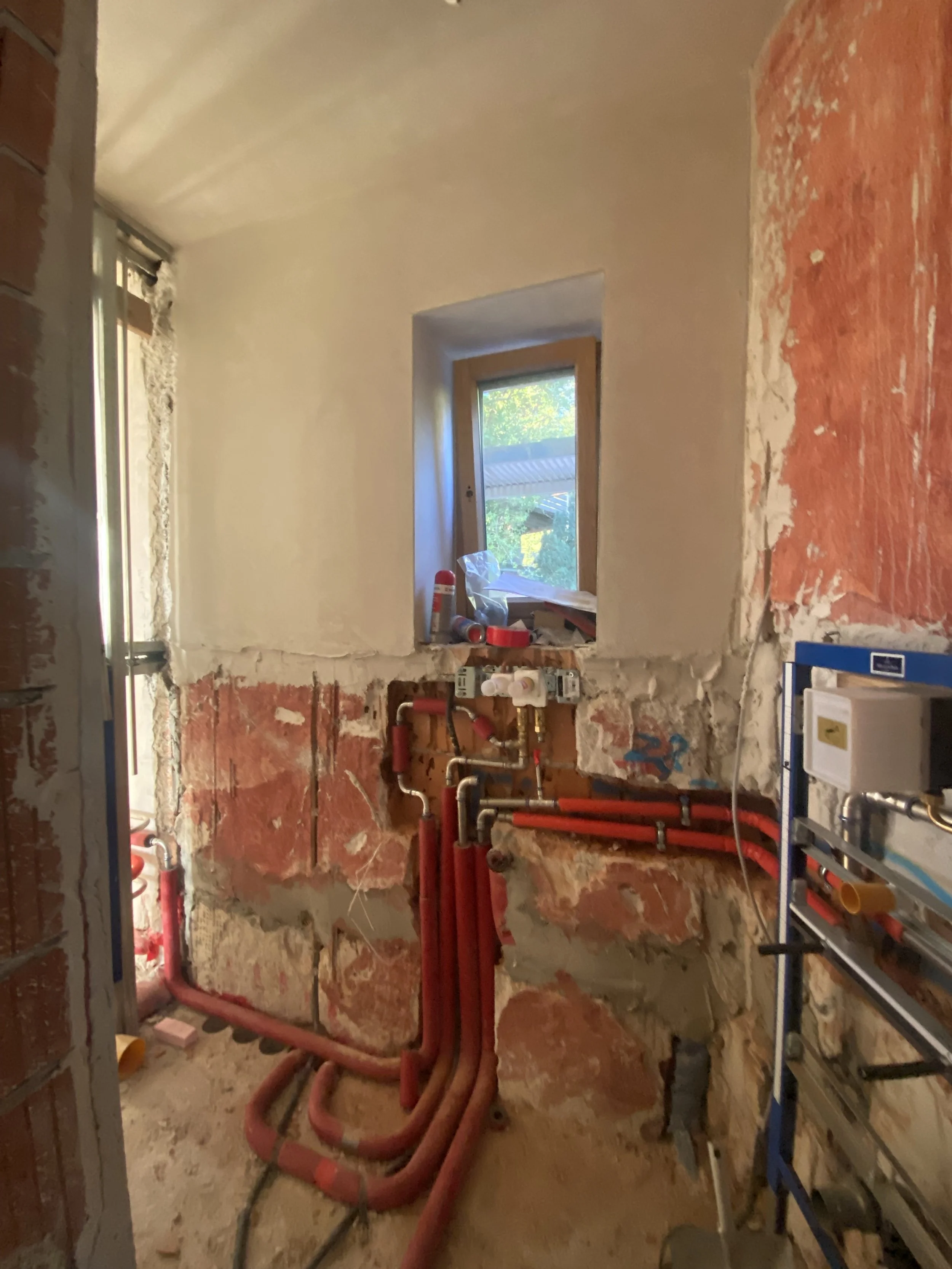 Innenraum einer renovierungsarbeiten Küche mit Wasserleitungen und einer kleinen Fensteröffnung.