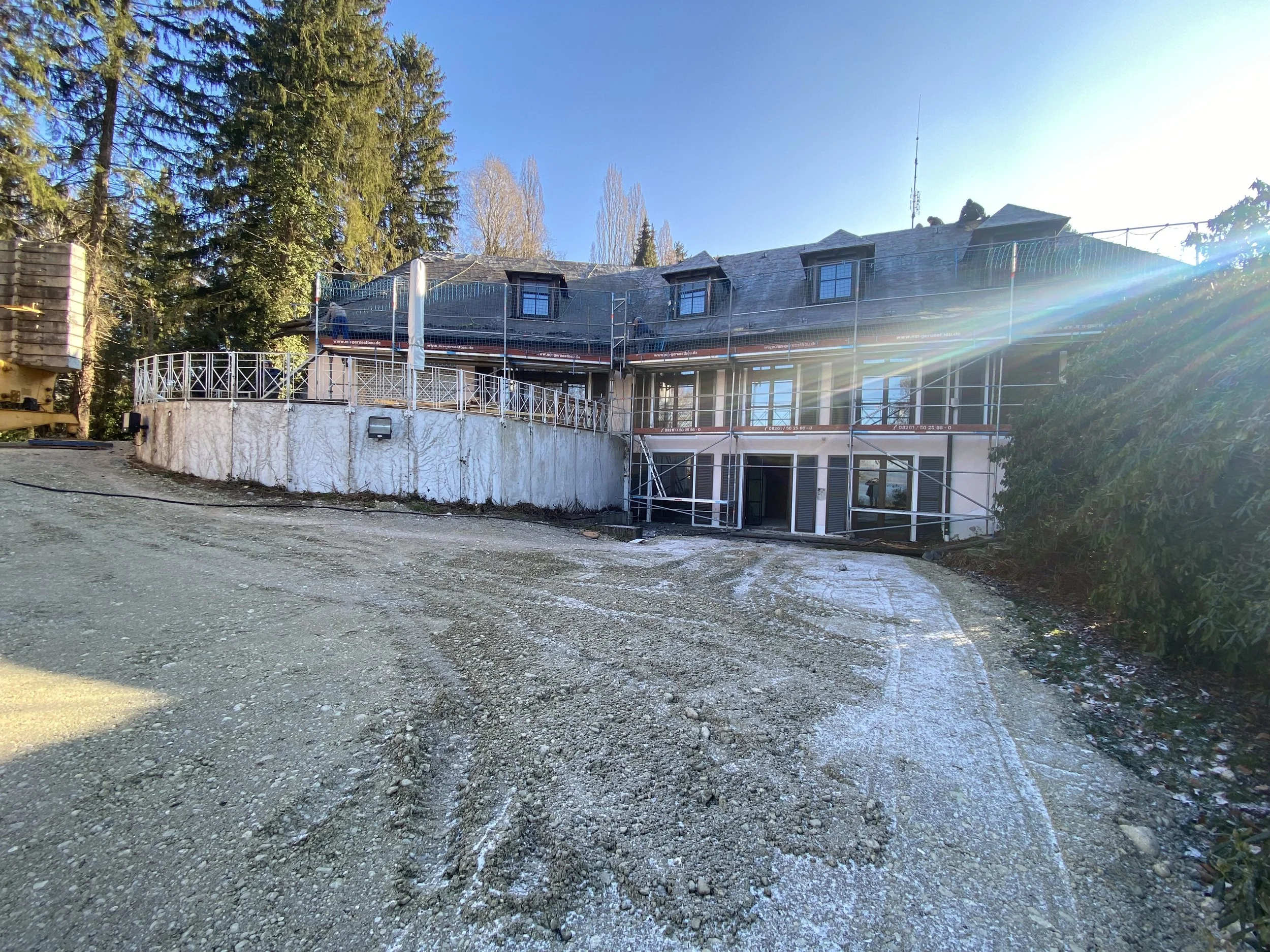 Ein mehrstöckiges Haus, das sich im Bautenschutz befindet, mit Bauzäunen und Gerüsten, umgeben von Tannenbäumen, bei sonnigem Wetter.