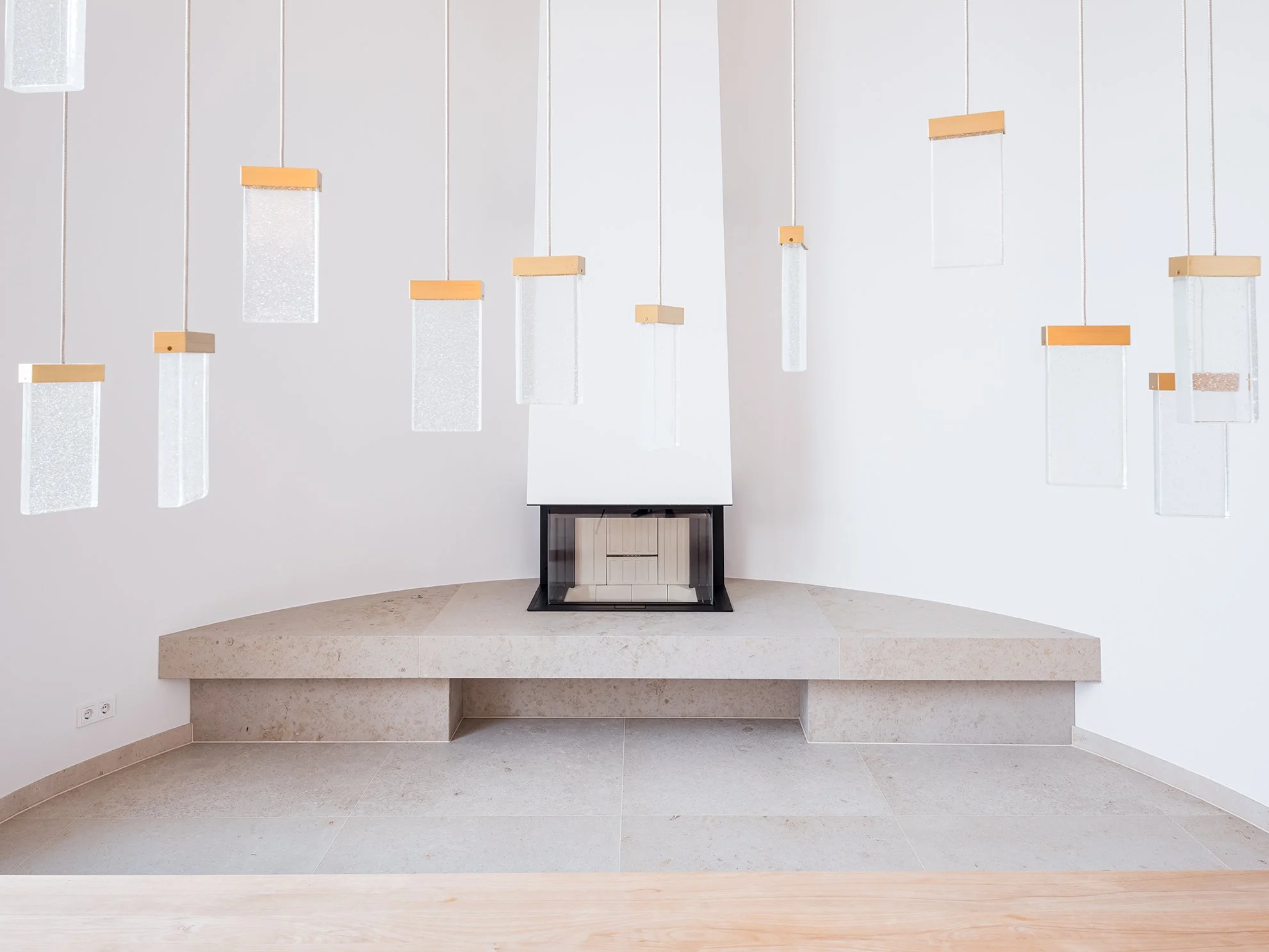 Ein moderner, schwarzer Kamin auf einem erhöhten, breiten Steinterrasse in einem minimalistischen Raum mit weißen Wänden und mehreren hängenden, rechteckigen Leuchten.