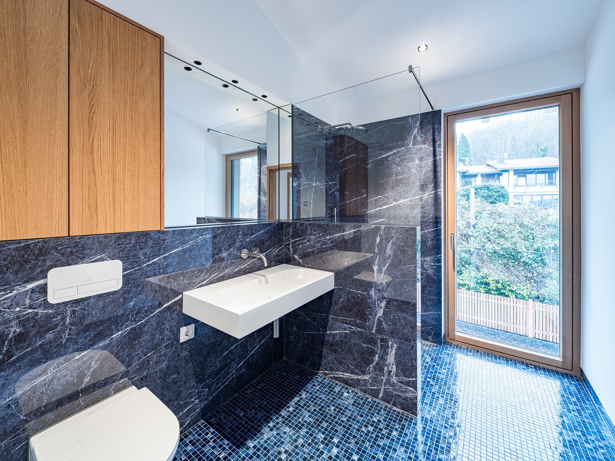 Moderner Badezimmer mit schwarzen Marmorfliesen, USB-gestütztem WC, weißer rechteckiger Handwaschbecken, großer Spiegel und Holzschränken, mit Fenster mit Blick auf eine grüne Landschaft.