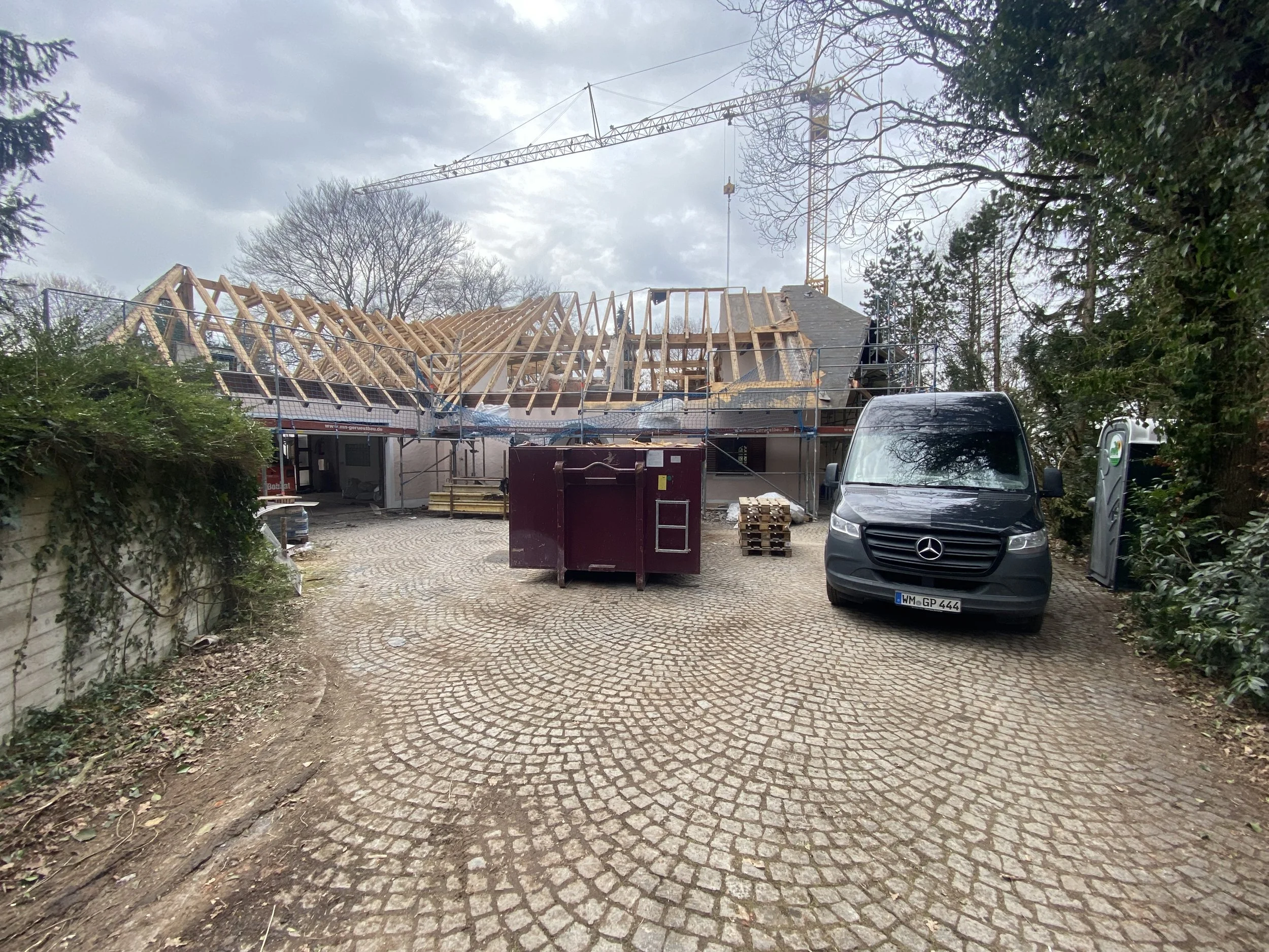 Ein Haus im Bau mit sichtbarer Holzkonstruktion auf dem Dach, Bauwagen, Baum und Baukran im Hintergrund, umgeben von Bäumen und Gebüschen, auf gepflastertem Hof mit Müllcontainer und Mercedes-Benz Transporter.