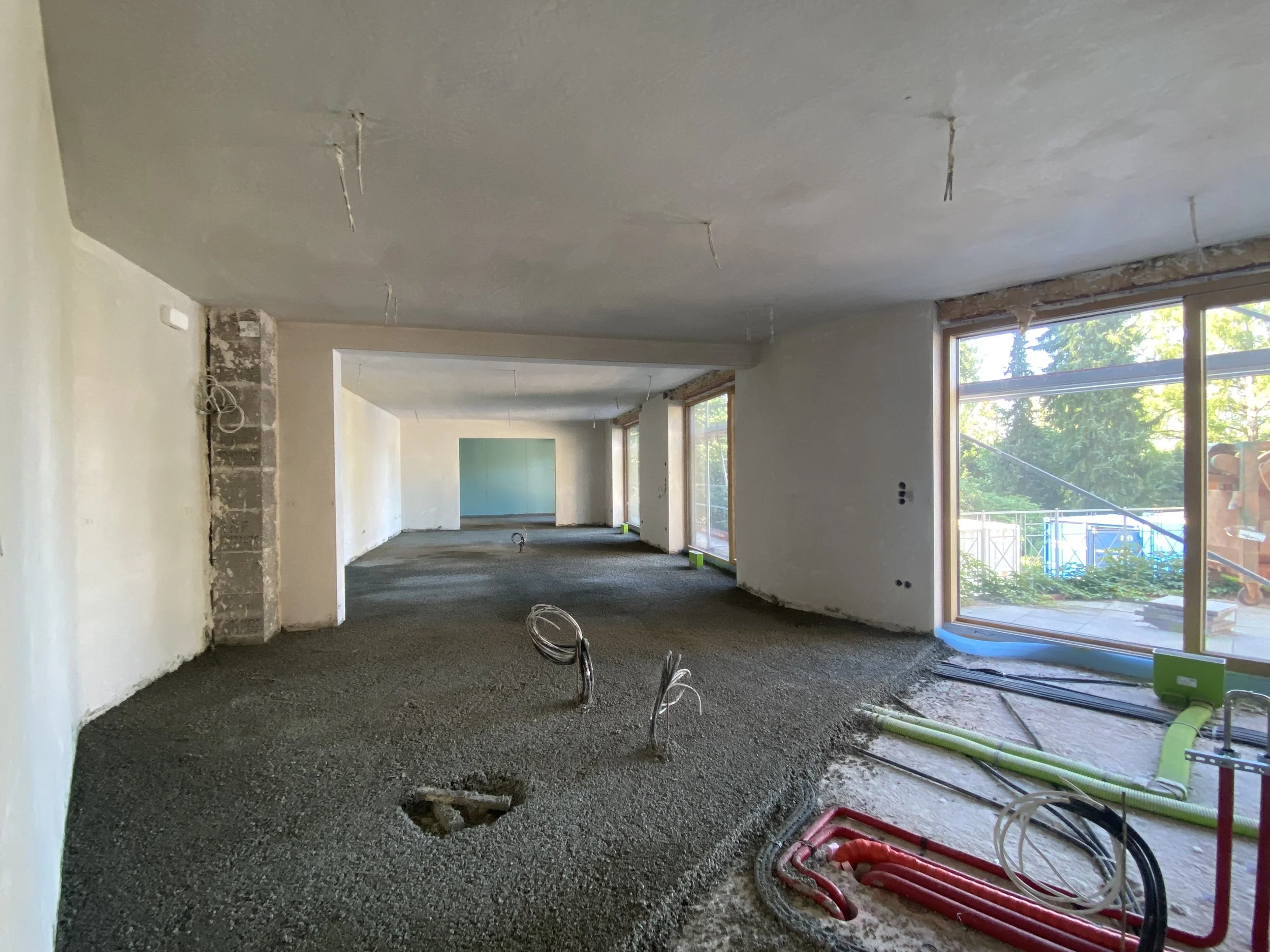 Innenraum eines im Bau befindlichen Gebäudes mit noch ungelegtem Boden und unvollendeter Wand- und Deckeninstallation, Fenster mit Blick ins Freie, sichtbare Rohrleitungen und Kabel.