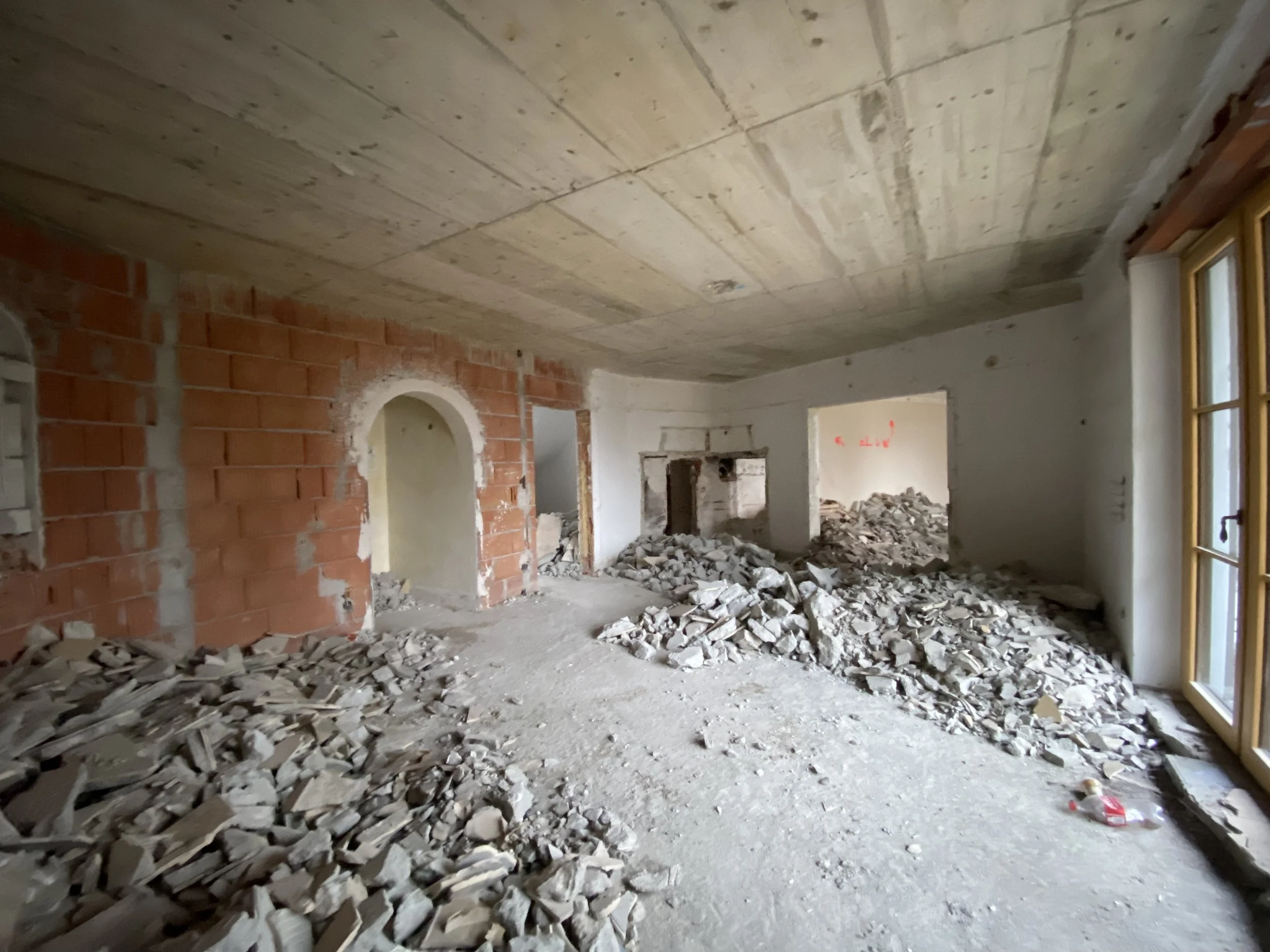Innenraum eines renovierungsbedürftigen Zimmers mit verstreutem Bauschutt, rotem Ziegelsteinwand und Fenstern