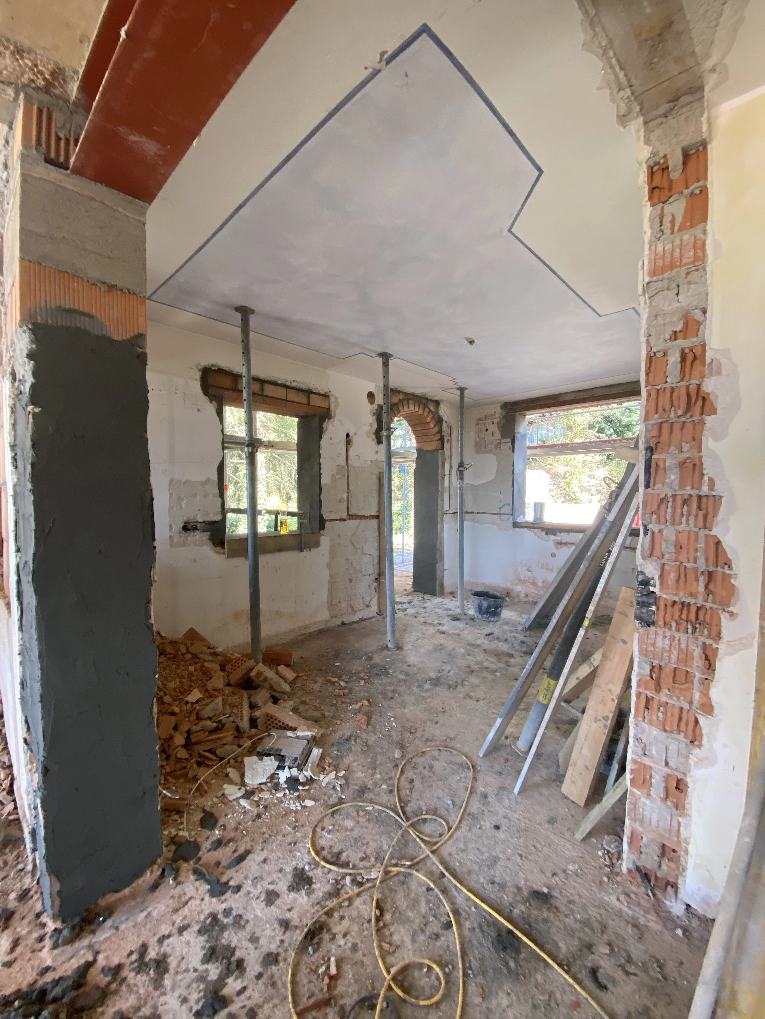 Innenraum einer Baustelle mit vorübergehend abgestützten Decke, Ziegelwänden und Fenstern, die noch unvollständig sind.