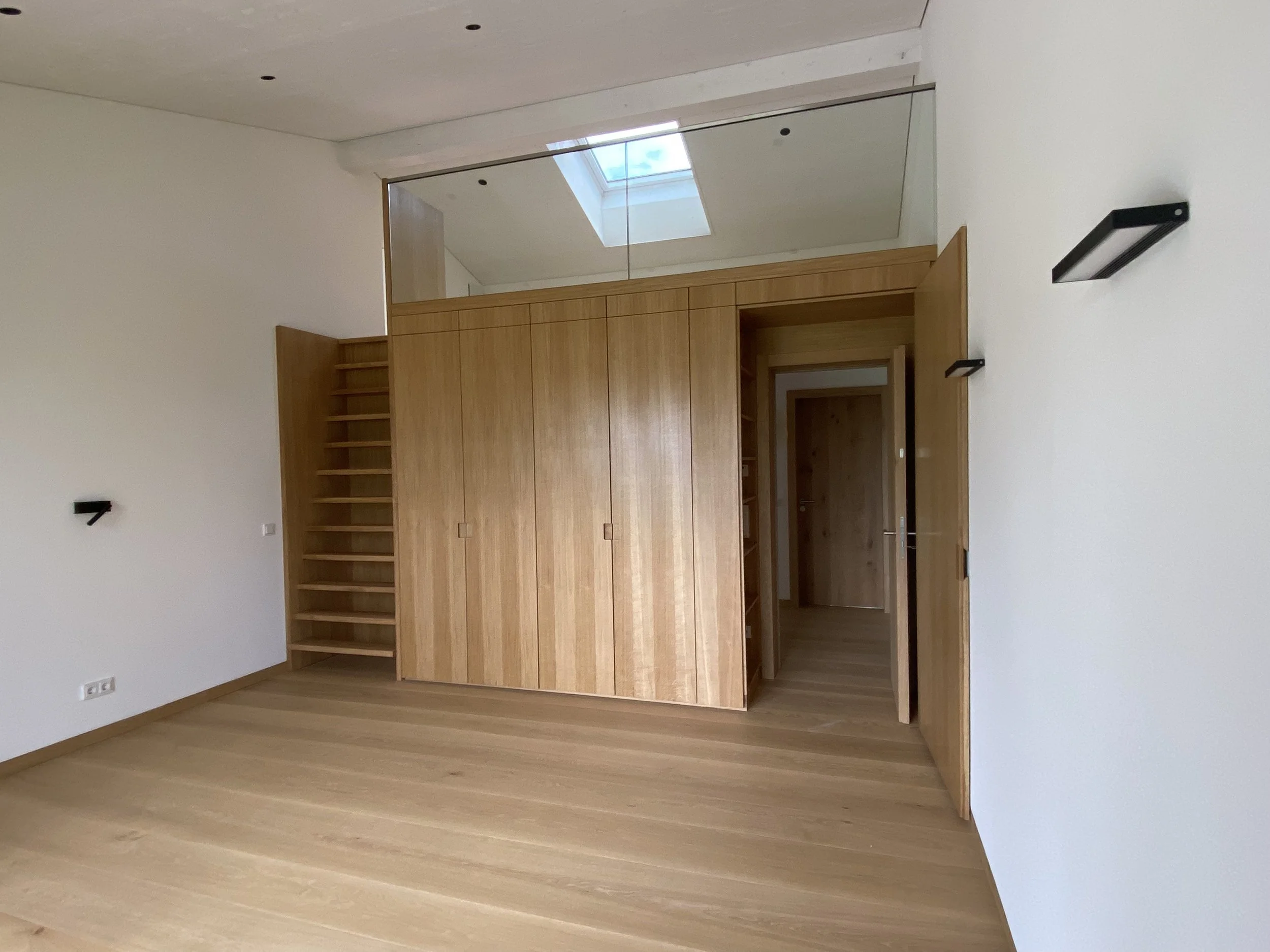 Ein leerer Raum mit Holzfußboden, weißen Wänden und einer Holztrennwand, die ein oberes Fenster und Regale enthält.