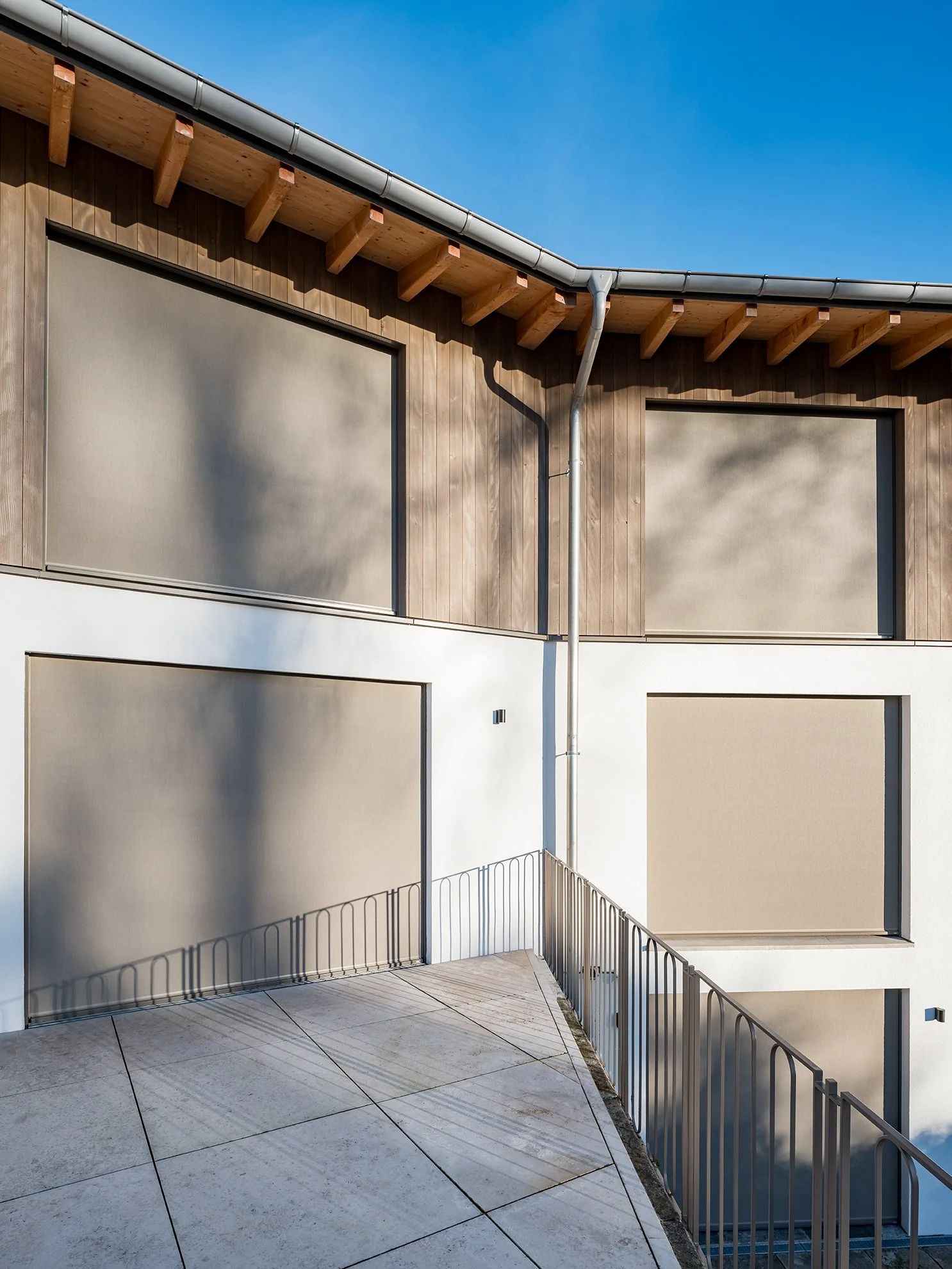 Moderne Gebäudefassade mit Holz- und Weißflächen, große Fenster mit Rollos, Balustrade, blauer Himmel, Schatten.
