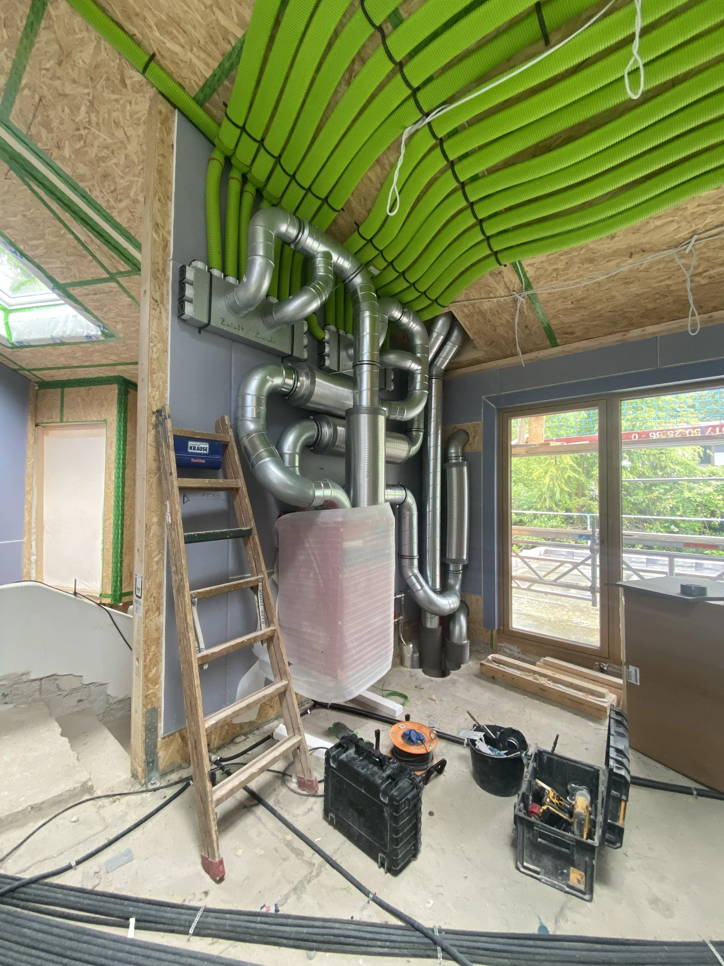 Bauarbeiten in einem Raum mit sichtbaren versilberten Lüftungsrohren, grünen Kabelschläuchen an der Decke, einer Leiter, Werkzeugkiste und großen Fenstern, durch die grüne Natur draußen sichtbar ist.