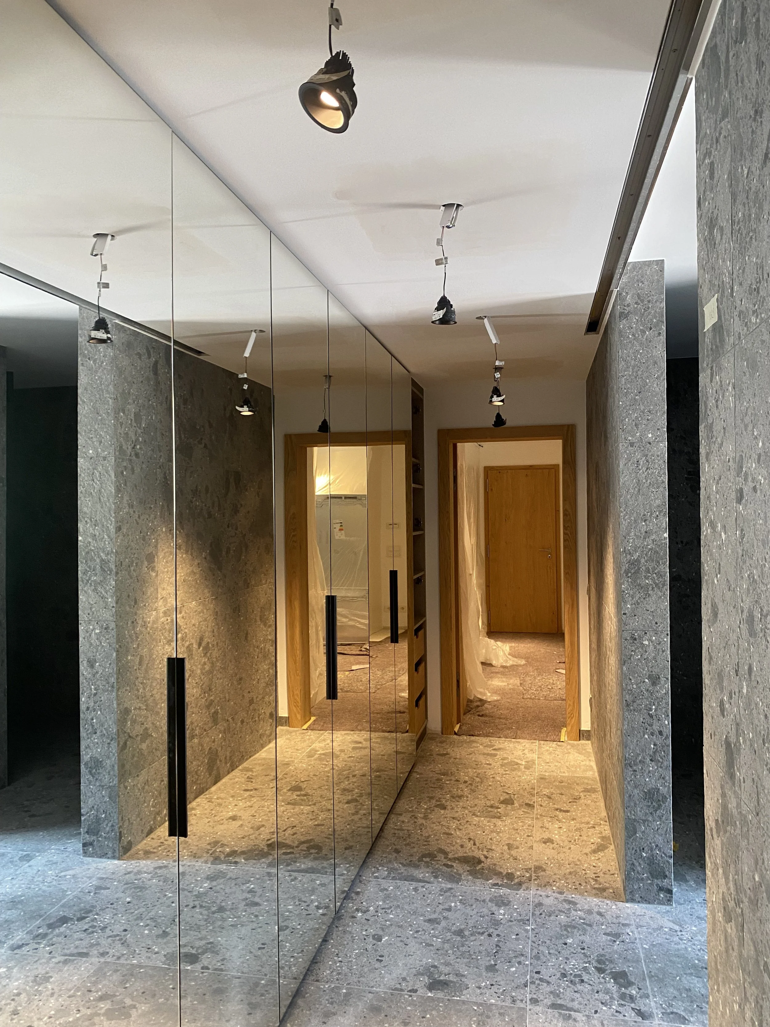 Innenansicht eines Flurs mit großen Spiegelflächen an der linken Wand, mehreren Deckenleuchten und Türen aus Holz, der Boden und die Wände sind aus grauem Stein.
