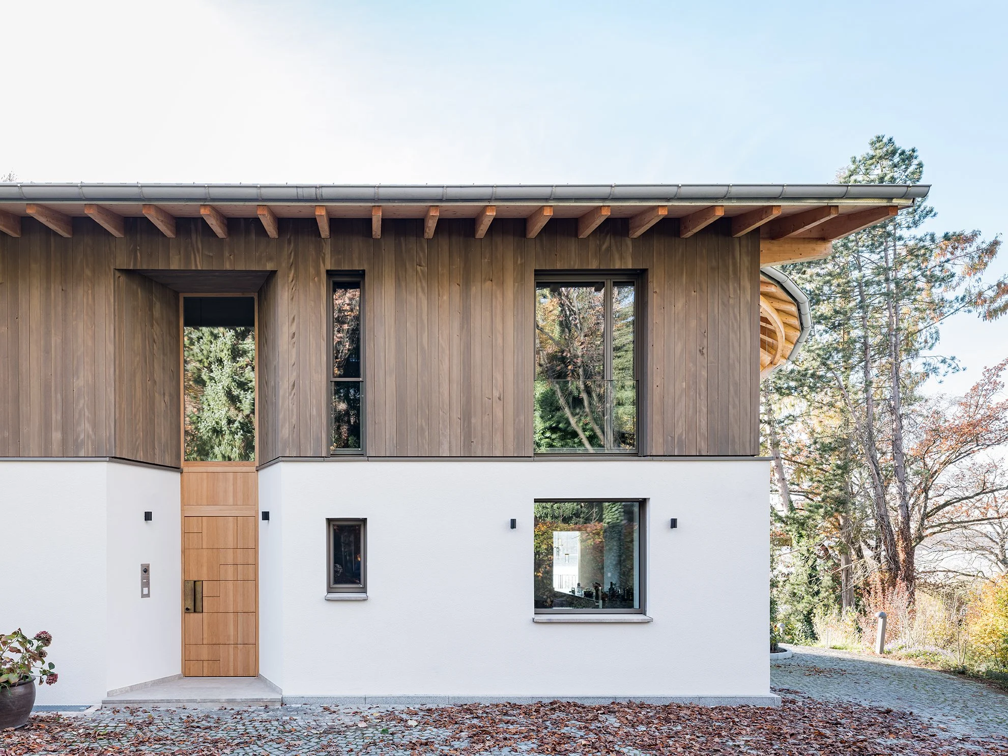 Modernes zweistöckiges Haus mit Holzfassade und weißen Wänden, umgeben von Bäumen und Herbstblättern.
