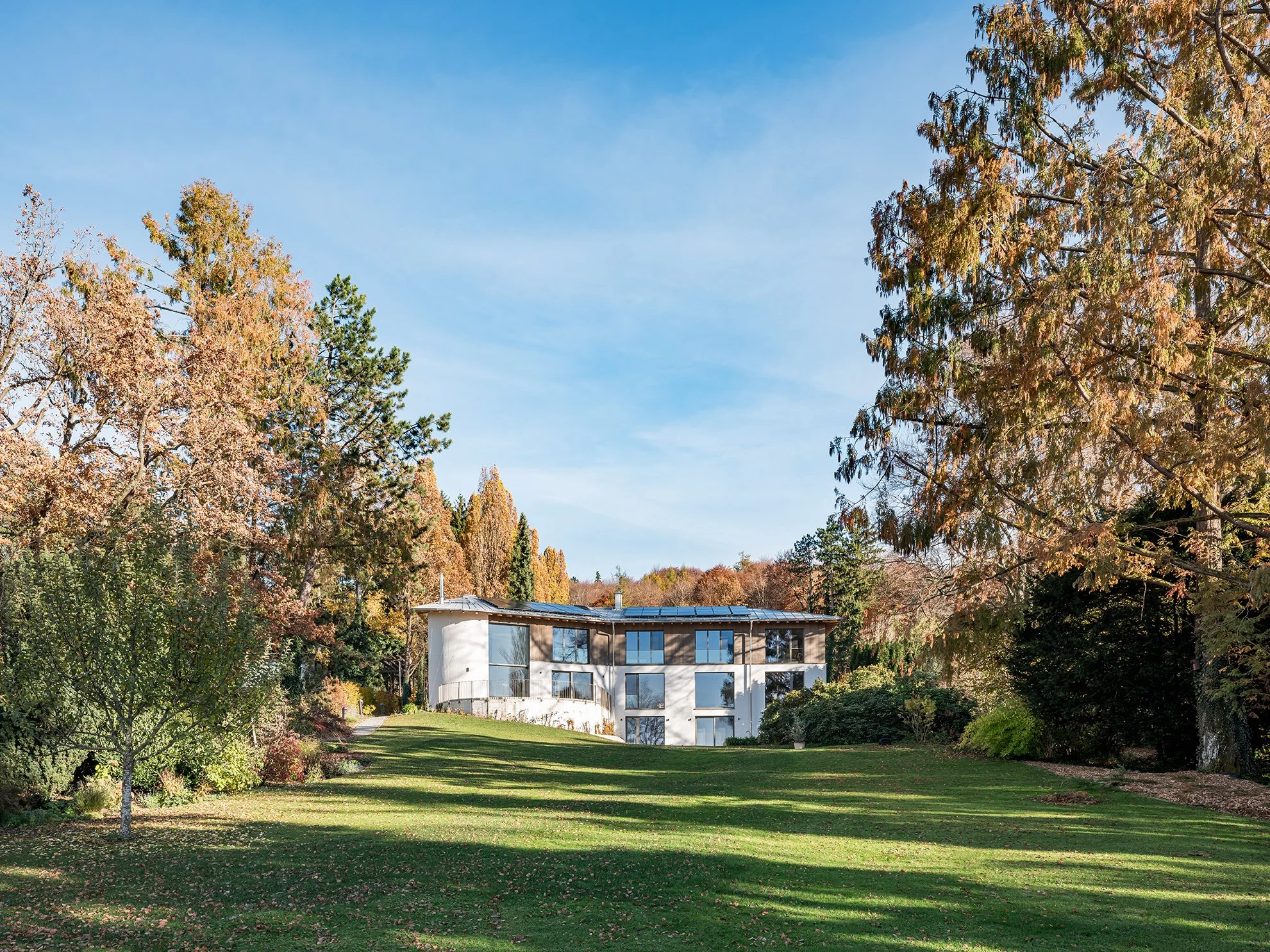 Modernes, mehrstöckiges Haus auf einem grünen, leicht ansteigenden Rasen mit Bäumen und Sträuchern, blauer Himmel, Herbstfarben.