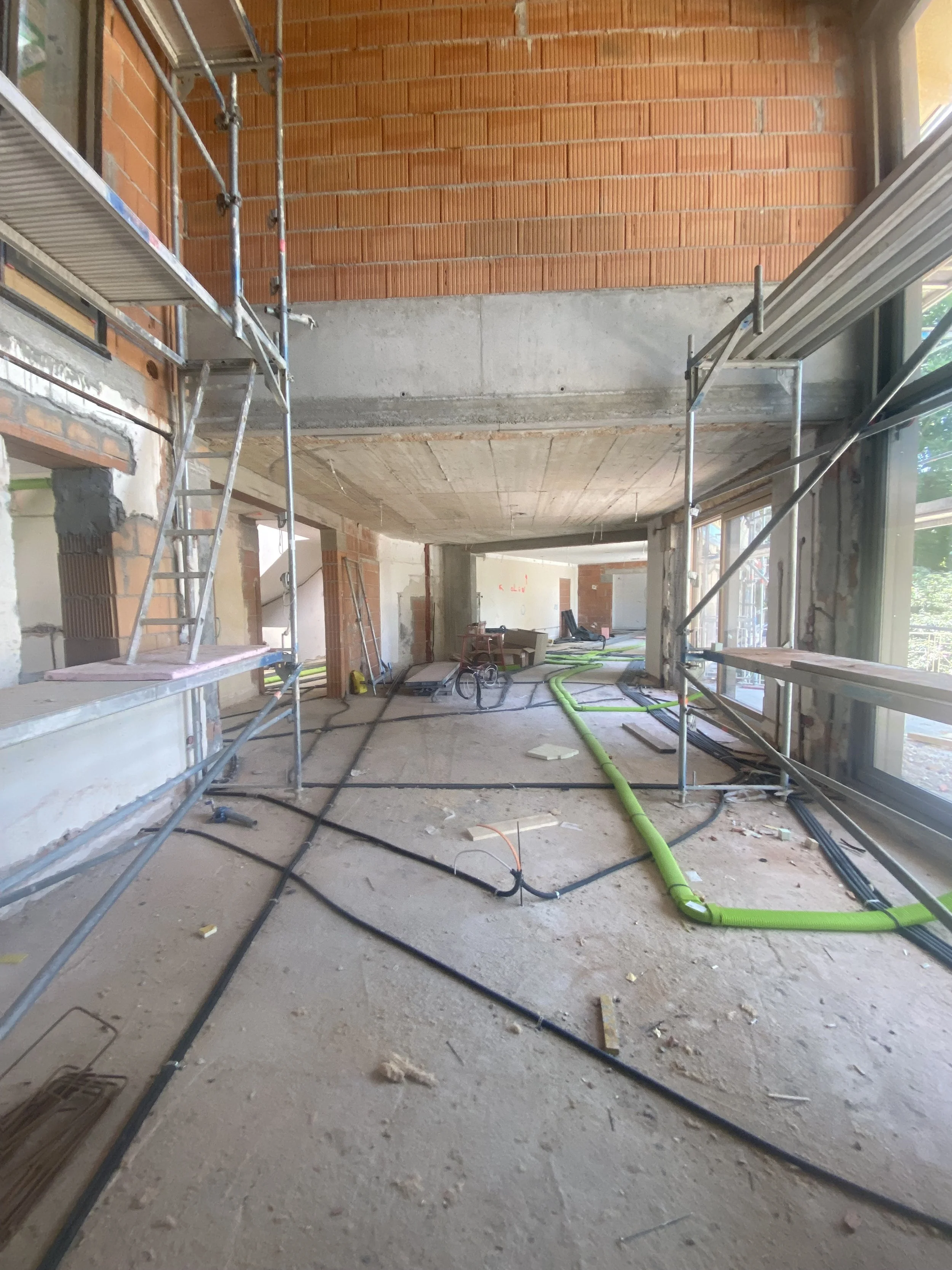 Innenraum eines Gebäudes unter Bau, mit sichtbaren Baugerüsten, Baufolie, Kabeln und Rohren auf dem Boden, große Fensterflächen, unvollendete Wände und Decken.