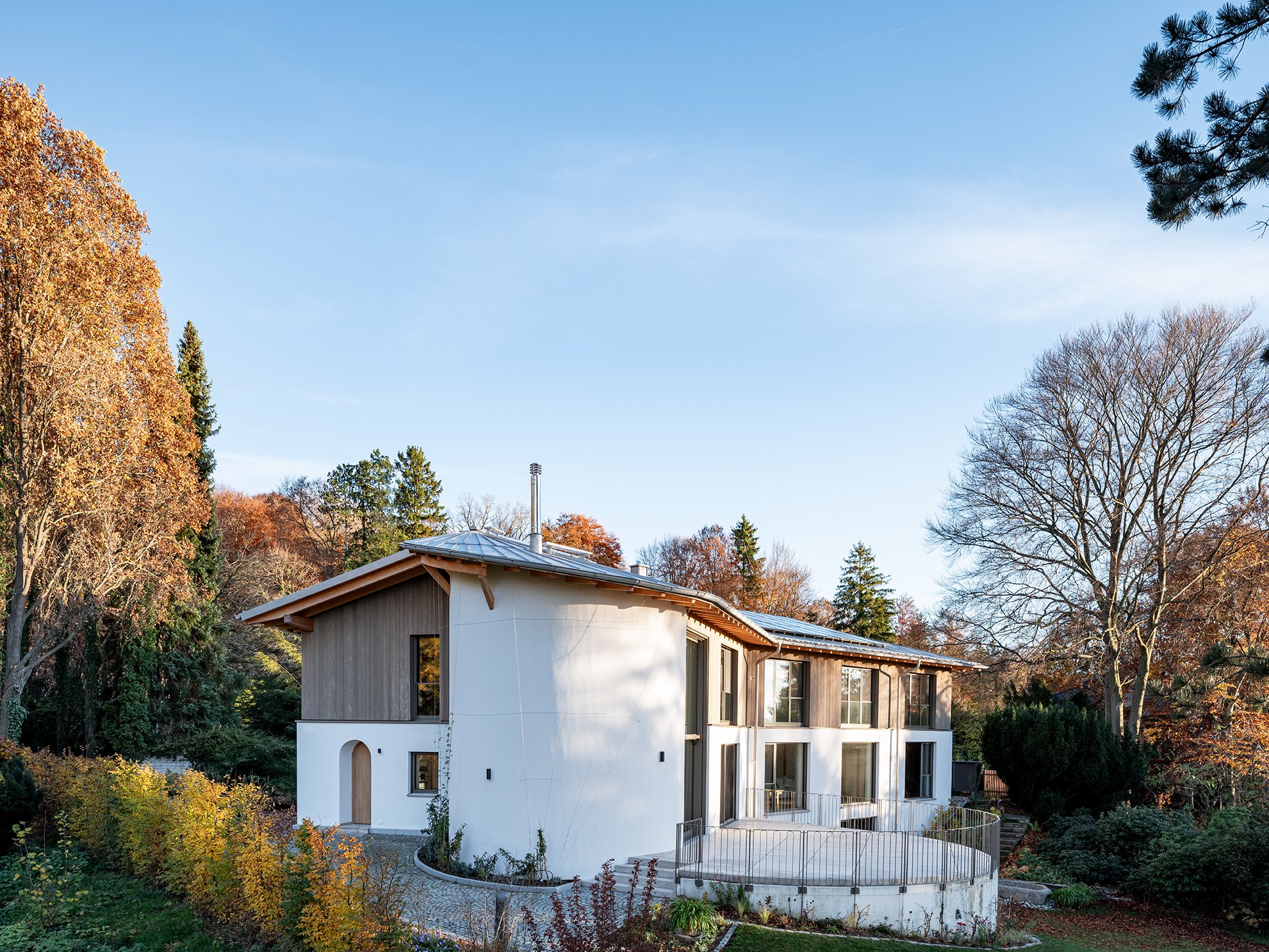 Ein modernes, zweistöckiges Haus mit weißen Wänden, Holzfassade und großen Fenstern, umgeben von Bäumen und einem Garten in herbstlicher Umgebung.