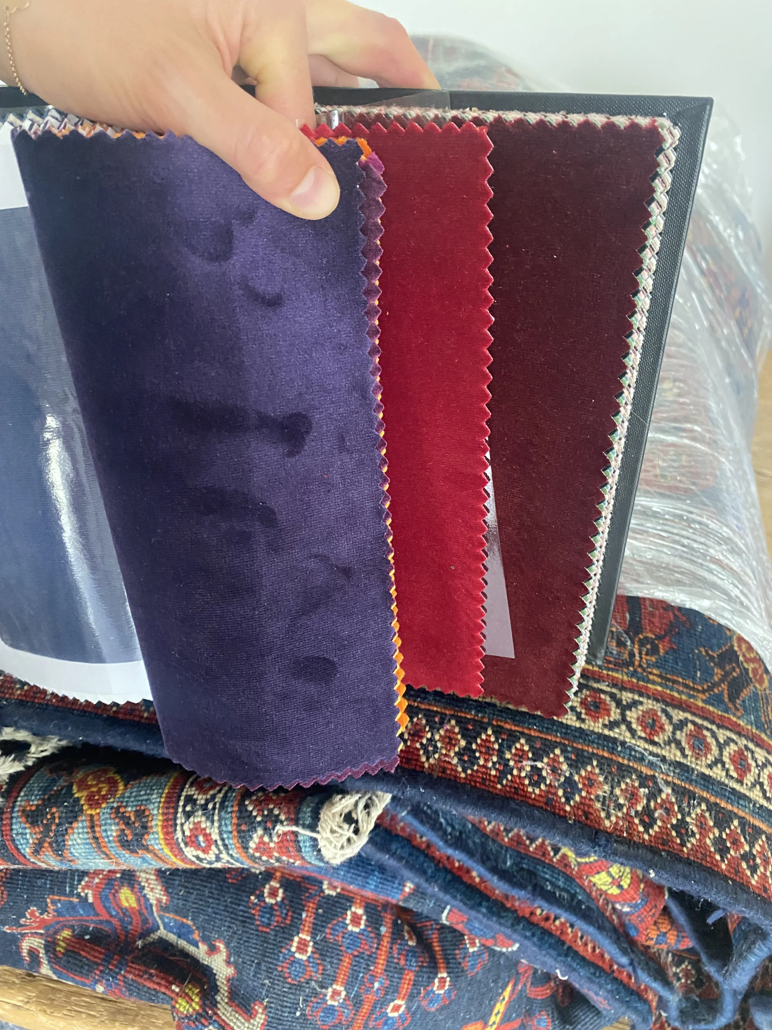 Farbige Stoffproben in Violett, Rot und Dunkelrot, gehalten von einer Hand, vor einem viktorianisch inspirierten Teppich mit Blumenmuster.