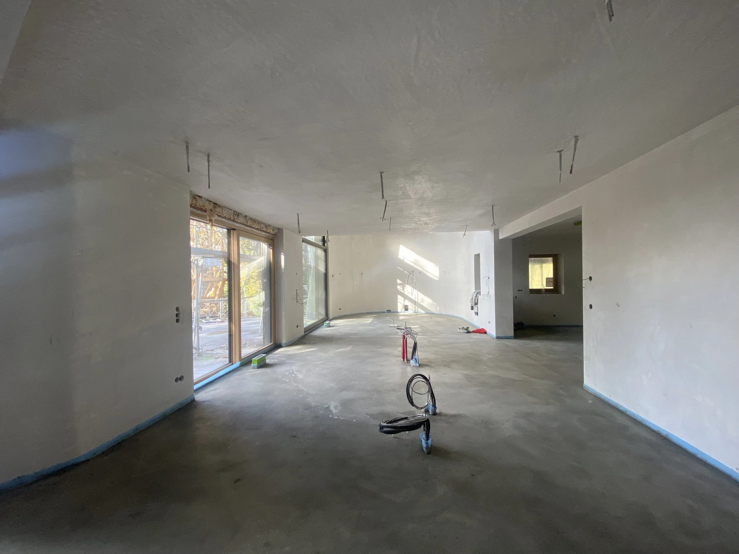 Innenansicht eines unfertigen Wohnzimmers mit großen Fenstern, unvollständiger Decke und Boden, sowie Installationsleitungen auf dem Boden.