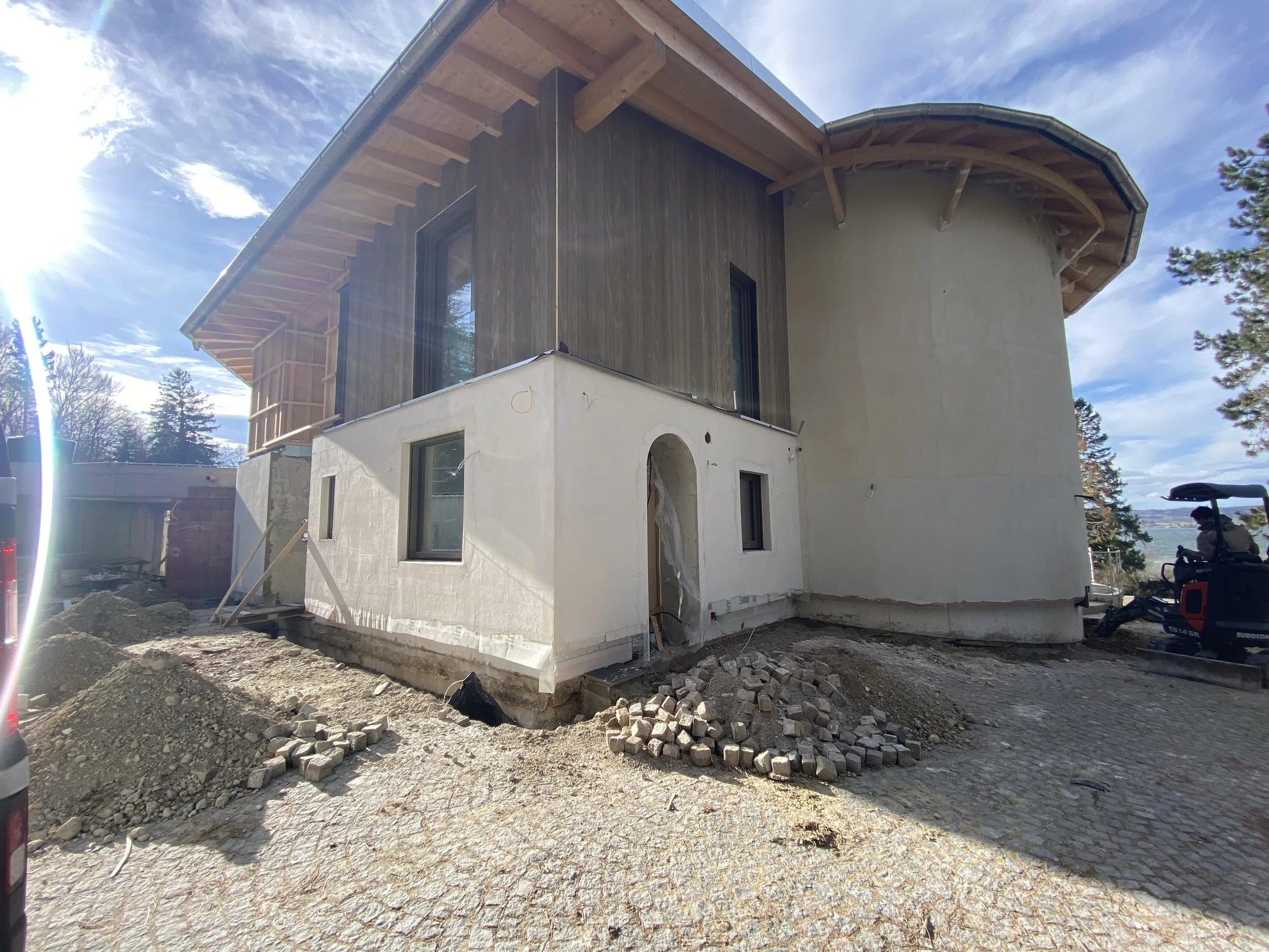 Baustelle eines modernen Hauses mit einer Kombination aus weißen und holzverkleideten Wänden, im Hintergrund ein blauer Himmel mit Wolken und Baustellenmaterialien im Vordergrund.