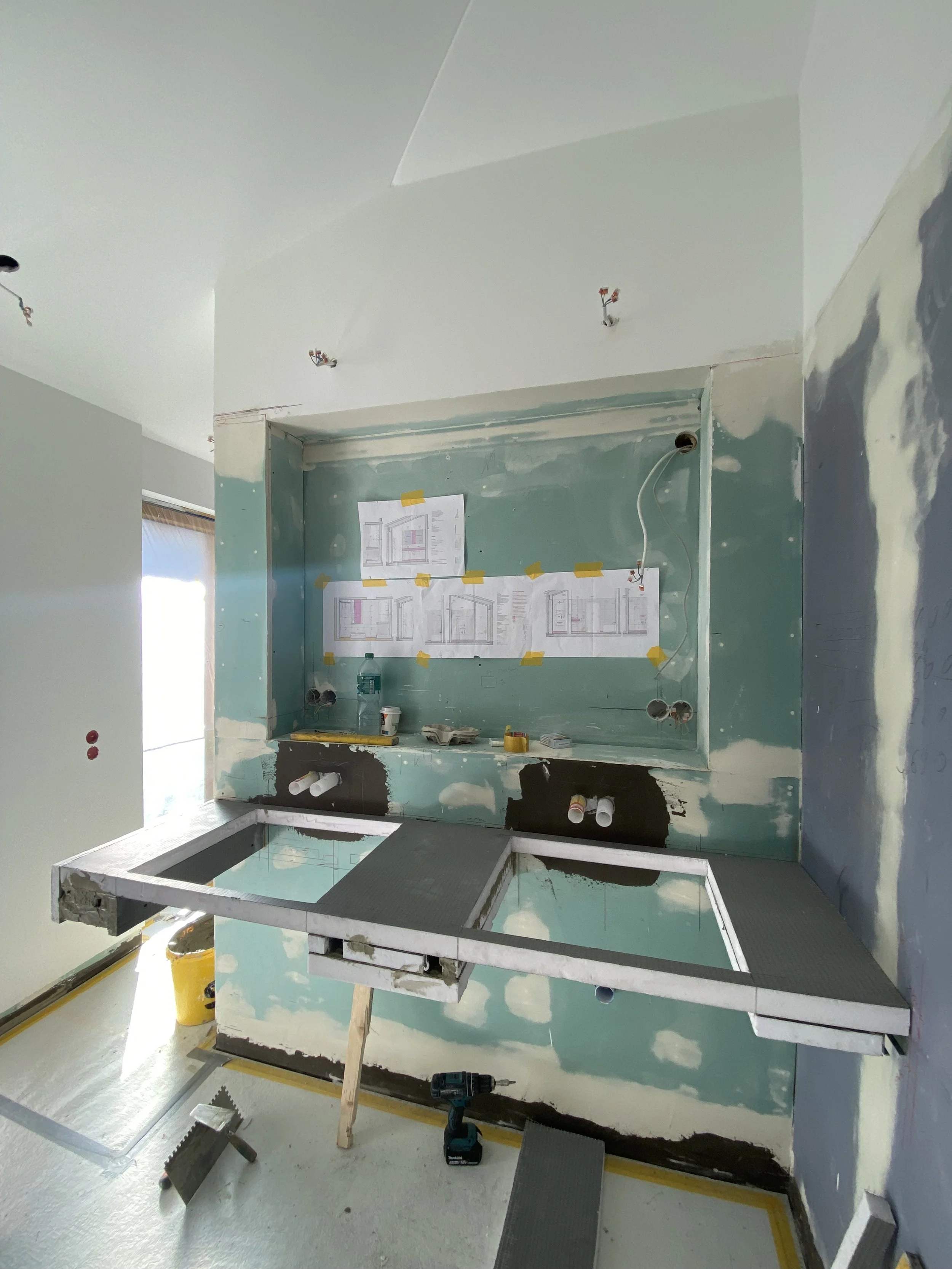 Badezimmer im Bau mit heraustretendem Waschbecken und Beleuchtung. Wände sind teilweise verspachtelt, mit Bauplänen an der Wand befestigt.