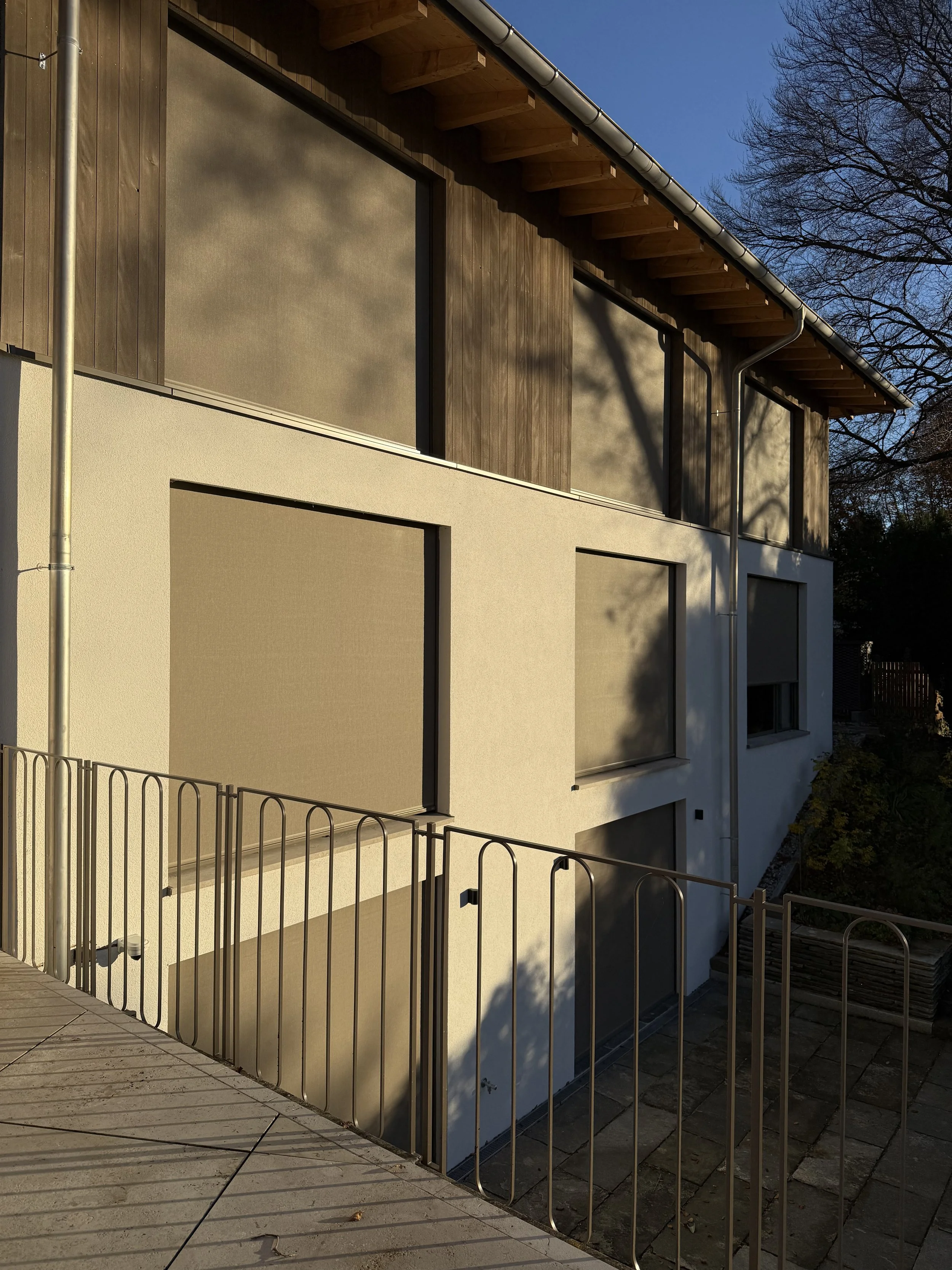 Moderne, zweistöckige Hausfassade mit Holz- und Putzflächen, Fenster mit Rollläden, umgeben von einem Garten mit Bäumen und einer Metallabsperrung.
