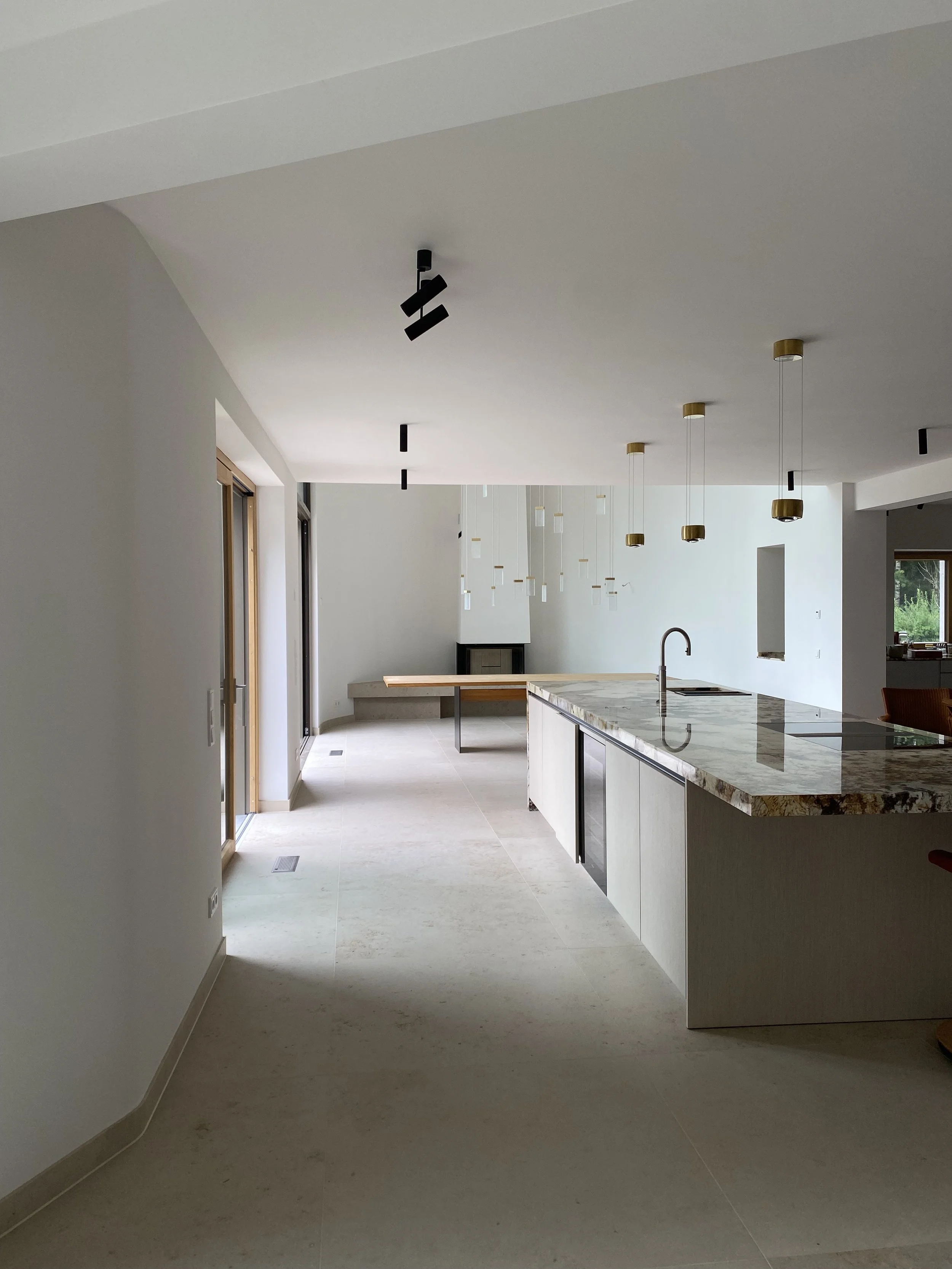 Moderner, offener Küchenraum mit weißen Wänden, einer Kücheninsel mit Granitoberfläche, Hängelampen, einem Kamin im Hintergrund und großen Fenstern, die Tageslicht hereinlassen.