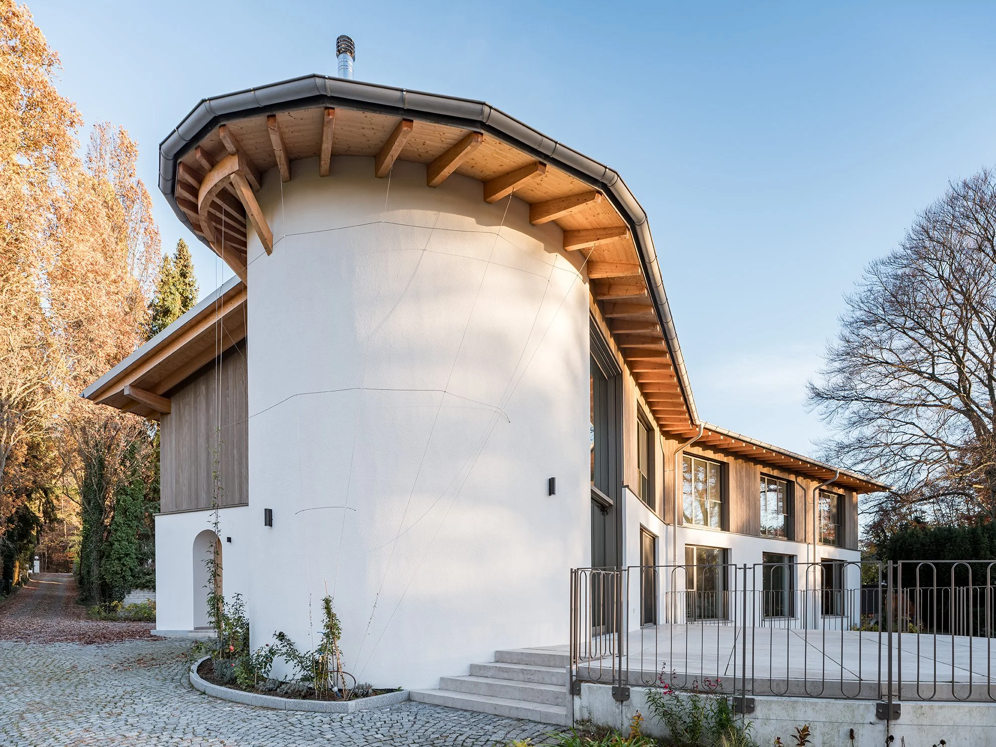 Modernes Haus mit weißen und holzverkleideten Fassaden, große Fenster, umlaufender Balkon, umgeben von Bäumen im Herbst, sonniges Wetter.