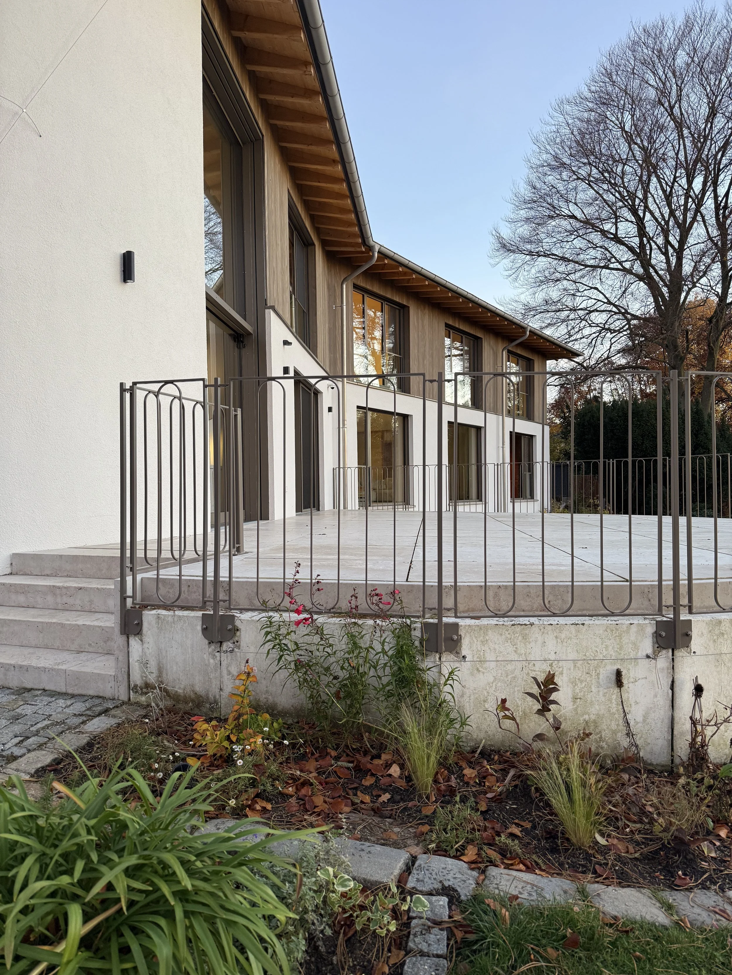 Modernes zweistöckiges Haus mit großen Fenstern, Holz- und Putzfassade, Terrasse mit Metallzaun und Garten mit Pflanzen und Blumen, im Hintergrund ein Baum ohne Blätter bei klarem Himmel.