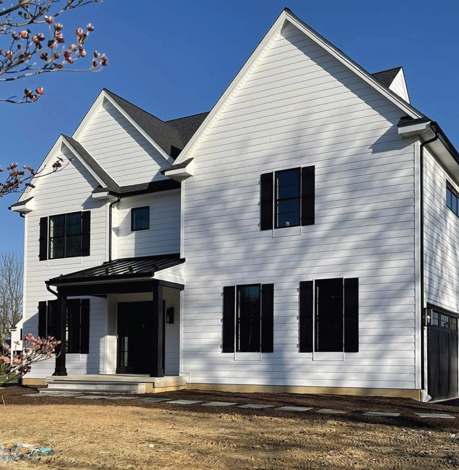 New white two-story house with black window shutters and a black front door, small front porch, and a blue sky background with a tree branch with pink flowers in the upper left corner.