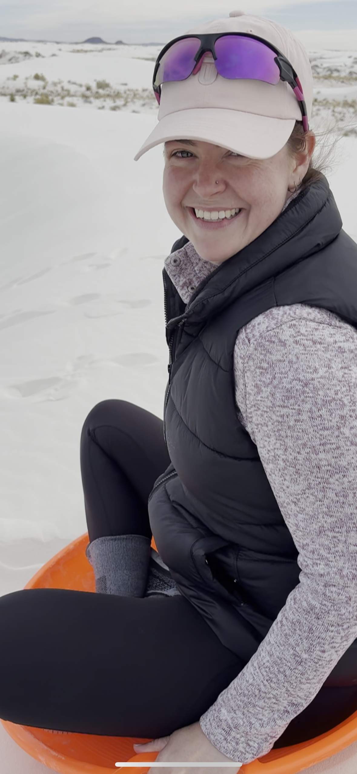 Woman smiling while sitting on an orange sled in a snowy landscape, wearing a beige cap with sunglasses, a black puffy vest, a pink and white patterned long sleeve shirt, and black pants.