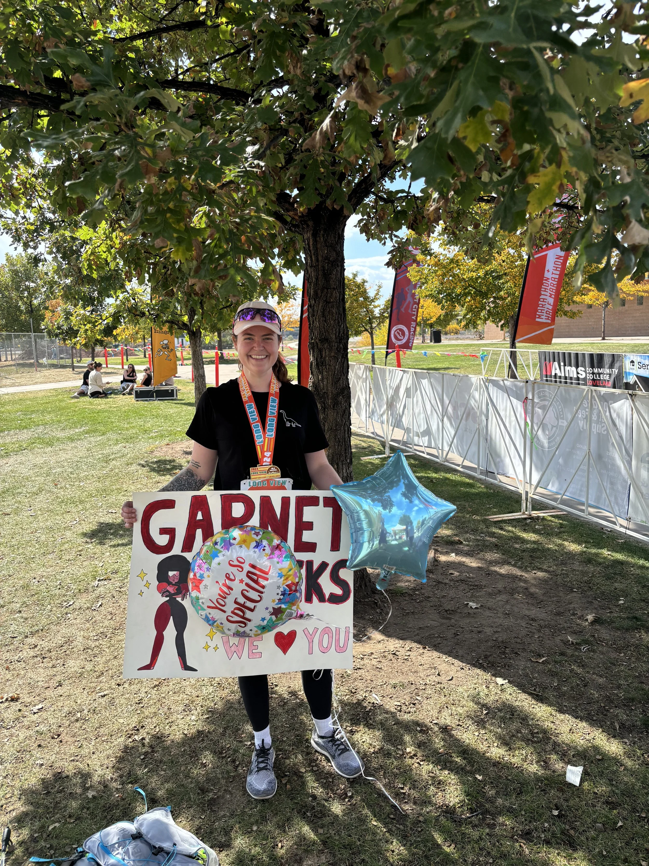 A woman smiling and holding a sign that says, "Garnet Rocks, You're So Special, We Love You" at an outdoor event, standing under a tree with balloons.