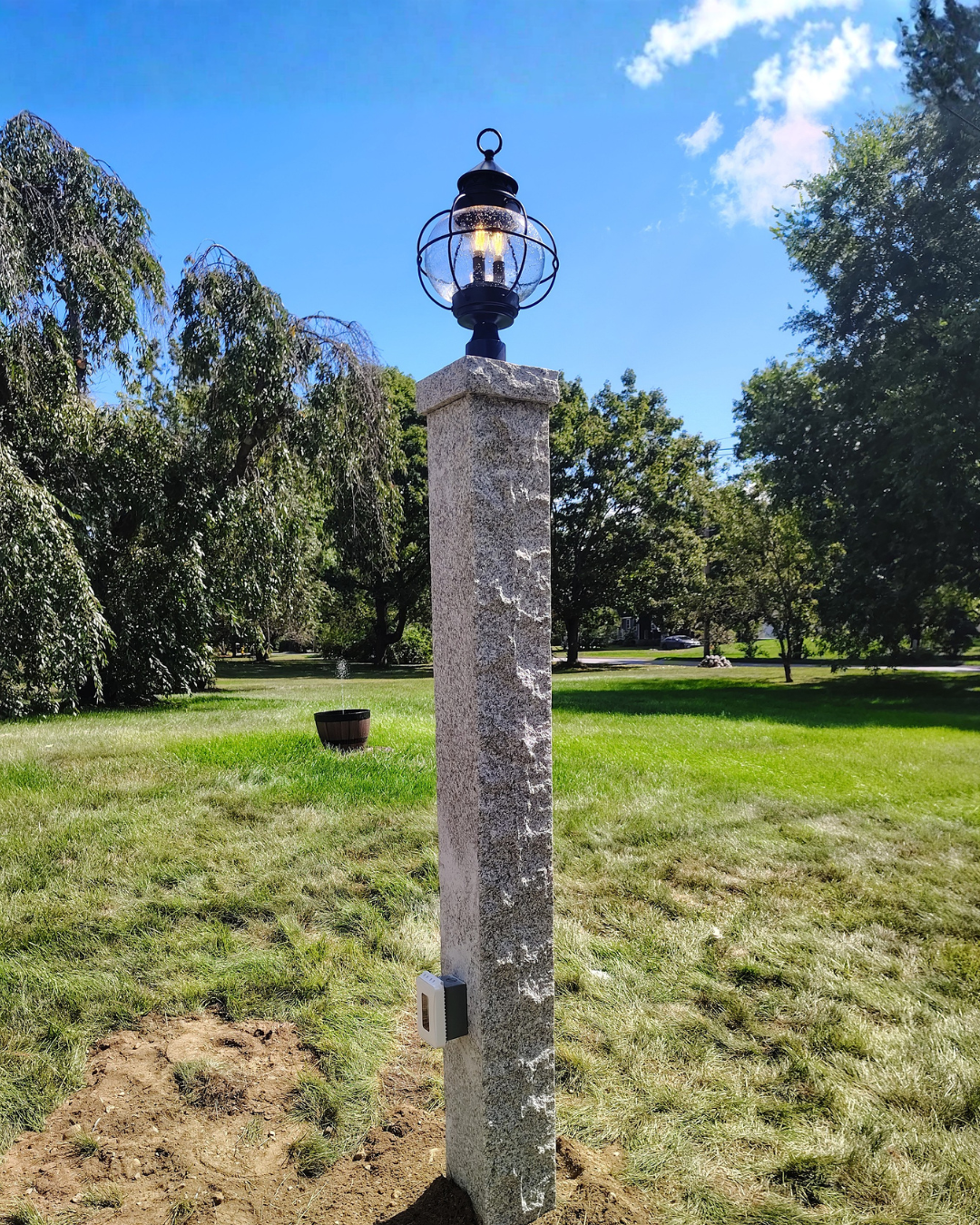 A stone lamp post with a black lantern on top, situated on a grassy lawn with trees in the background and a clear blue sky.
