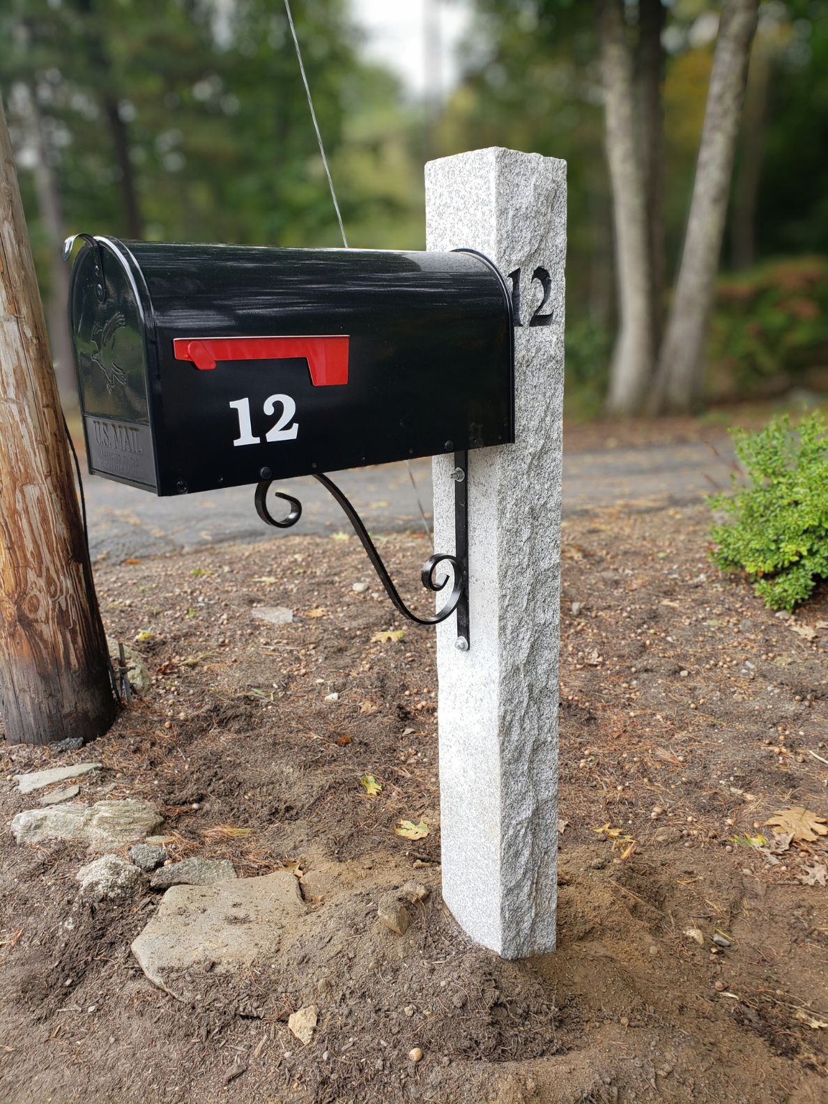 Black mailbox with red flag, marked with the number 12, mounted on a stone post next to an electrical pole on a dirt ground nearby trees.