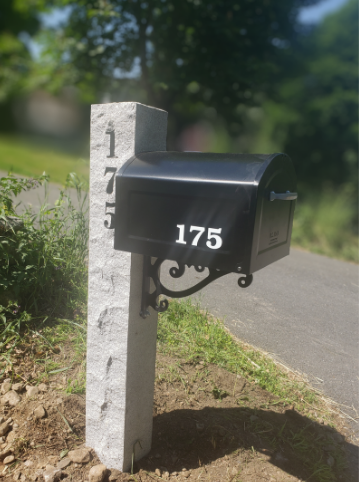 Black mailbox with number 175 on its front, mounted on a stone post with numbers 175, 177, 179 along the side, next to a sidewalk and green lawn with trees in the background.