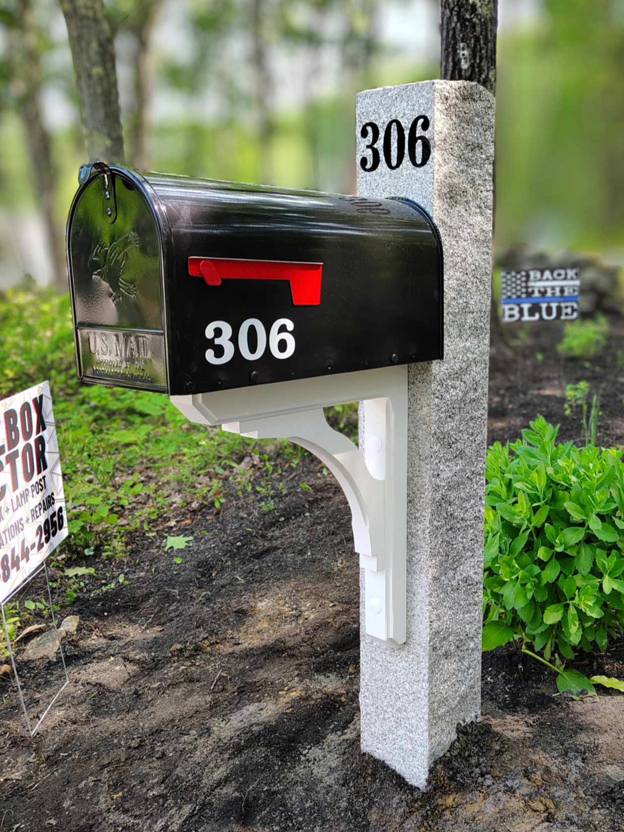 A black mailbox with the number 306 on it, mounted on a white post next to a concrete marker with the number 306, in a garden with mulch, green plants, and trees in the background.