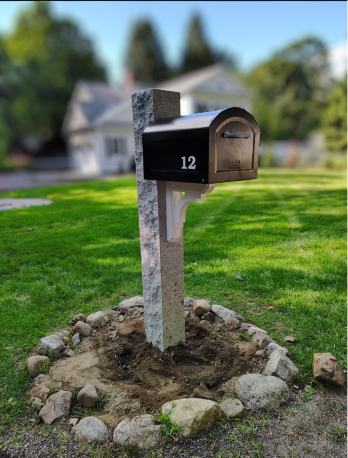 Black mailbox with the number 12 on a white post, set in a landscaped yard with green grass and a large house in the background.