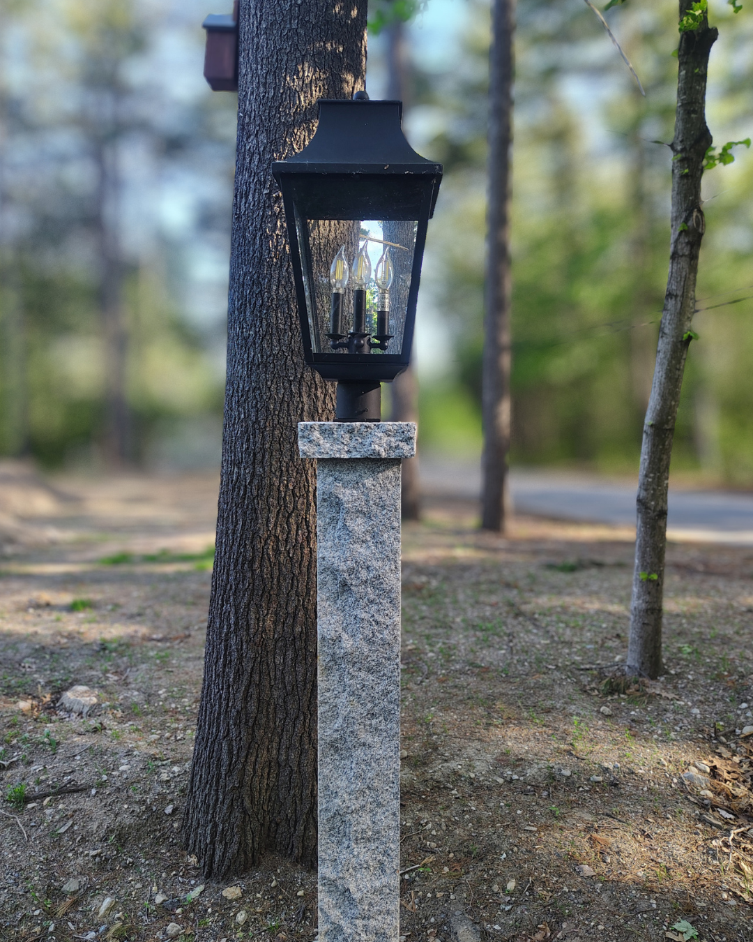 An outdoor black lantern-style light fixture with three candle-shaped bulbs, mounted on a gray stone post next to a tree in a wooded area.