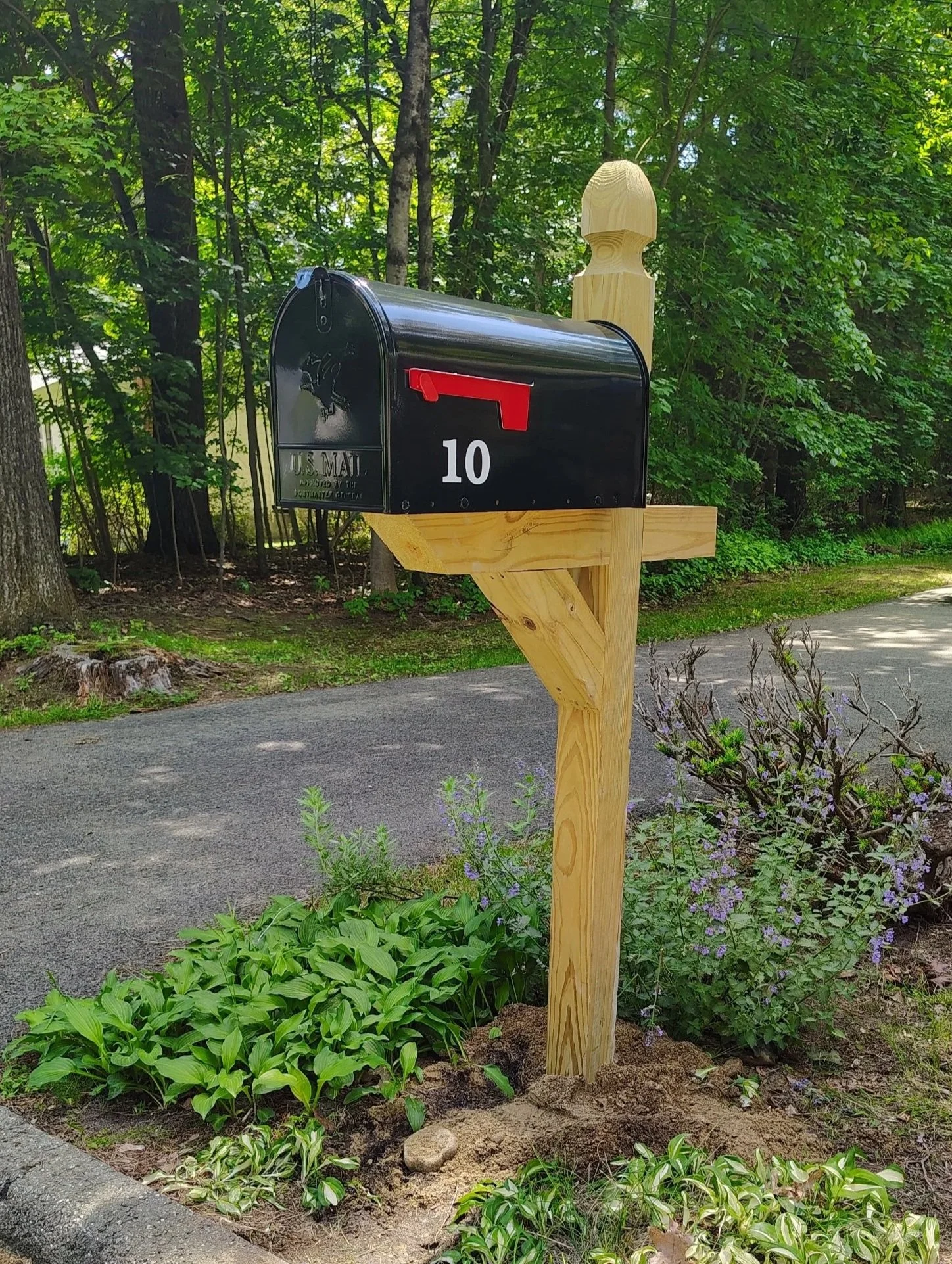 A black USPS mailbox with a red flag mounted on a wooden post in a garden with green foliage, purple flowers, and a paved pathway.