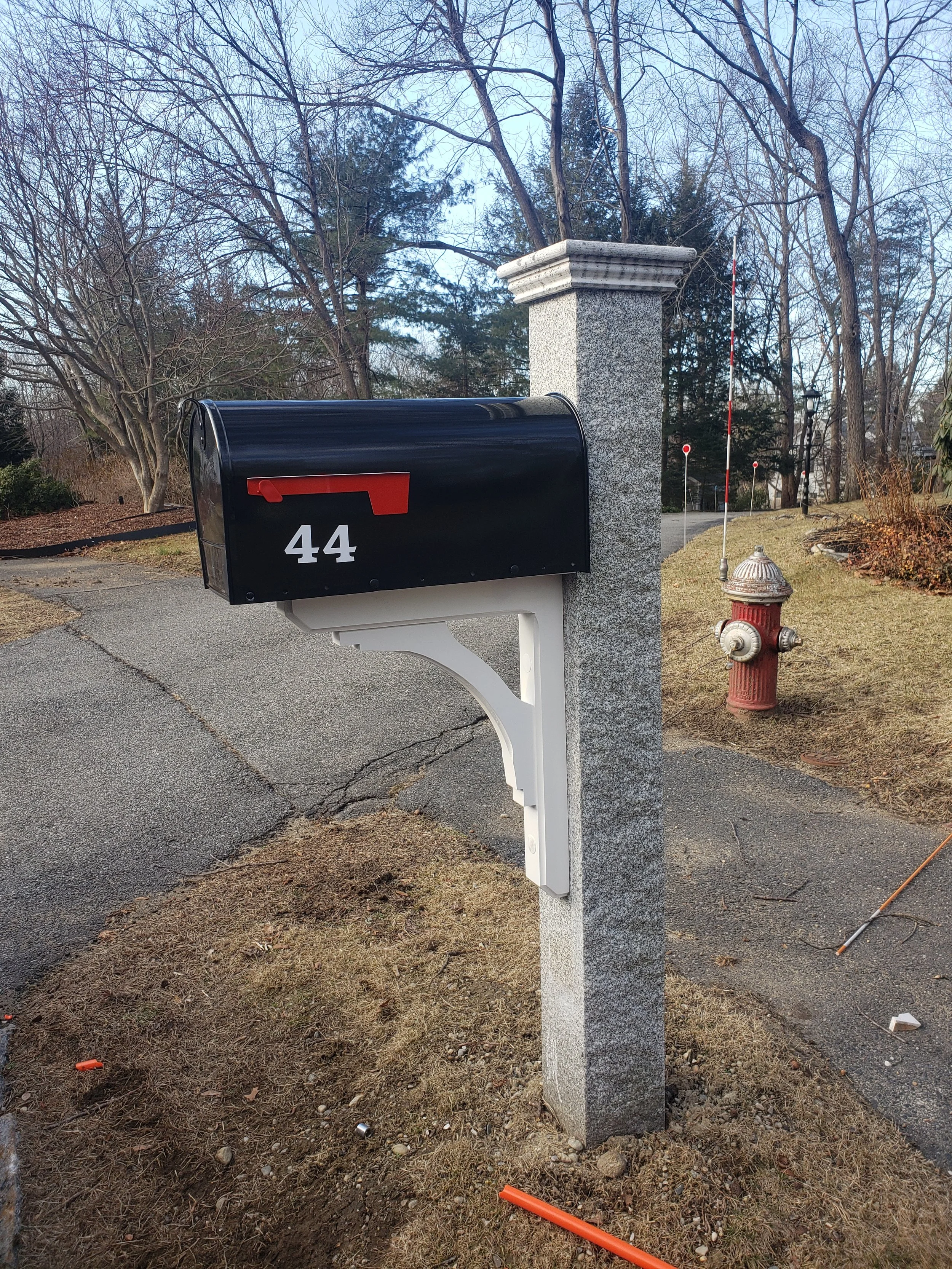 A black mailbox with the number 44 in white, mounted on a decorative white post next to a gray stone column, with leafless trees and a fire hydrant in the background.