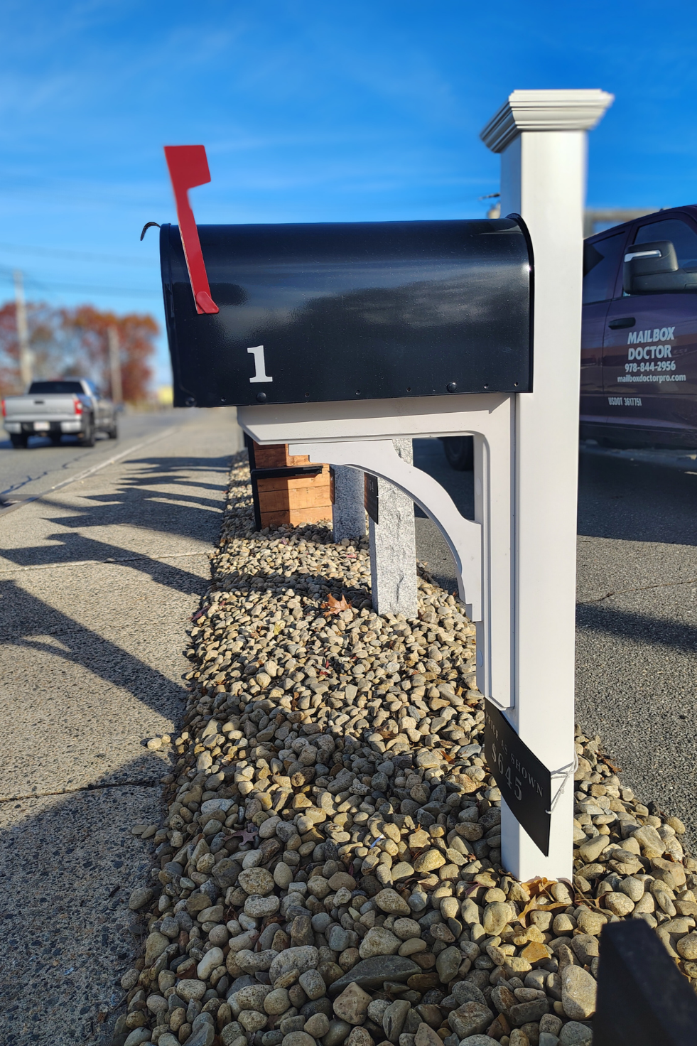 Black mailbox with red flag mounted on a white post, numbered 1, along a sidewalk with pebbled ground, under blue sky with some vehicles in the background.
