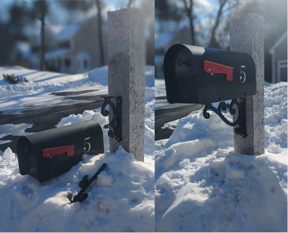 A black mailbox with the number 5 and a red flag, knocked over in the snow, in front of a concrete mailbox post, with a house and snow-covered ground in the background.