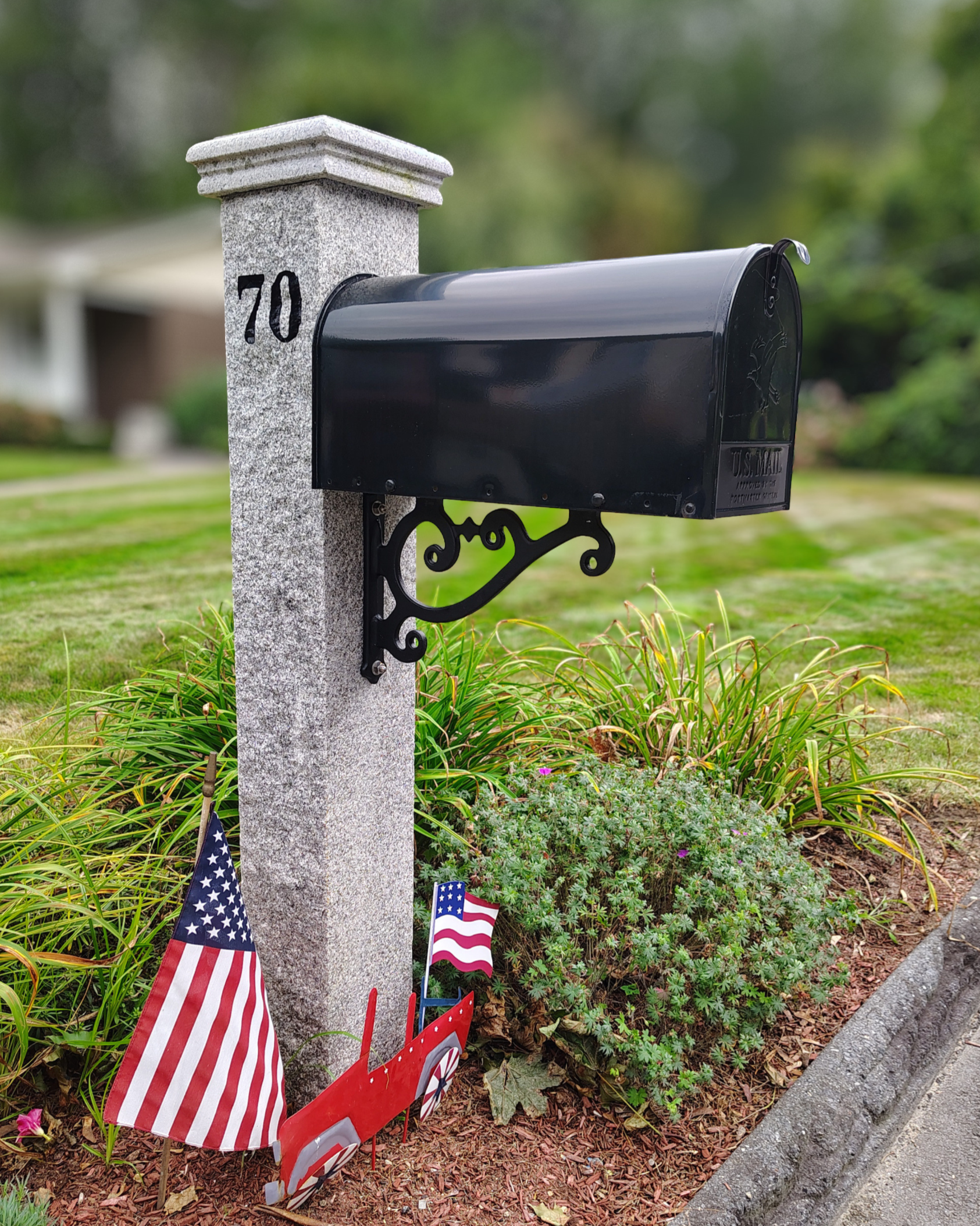 A black mailbox attached to a gray stone pillar with the number '70' painted on it. Small American flags are placed in the garden bed below the mailbox, which is bordered by a gray stone edge and green plants with purple flowers.