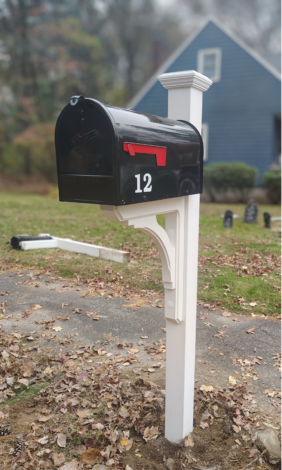 A black mailbox with the number 12, mounted on a white post in a suburban yard, with a house and fallen leaves in the background.