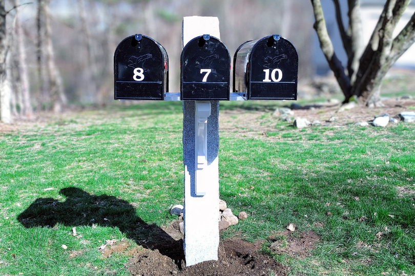 Three black outdoor mailboxes labeled 8, 7, and 10 mounted on a white post in a grassy yard.
