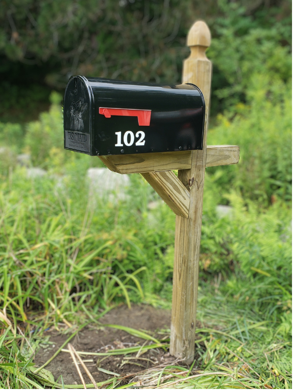 Black mailbox with the number 102 on it mounted on a wooden post in a grassy area with green foliage in the background.