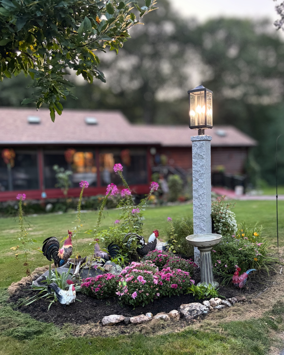 A garden bed with pink flowers, decorative rooster and hen statues, a stone bird bath, and a tall lamp post with a lit lantern, in front of a house with a red roof and a covered porch.