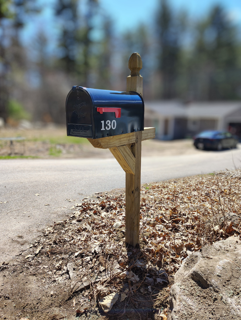 Front view of a black mailbox with the number 130 on it, mounted on a wooden post near a residential road, with blurred trees and houses in the background.
