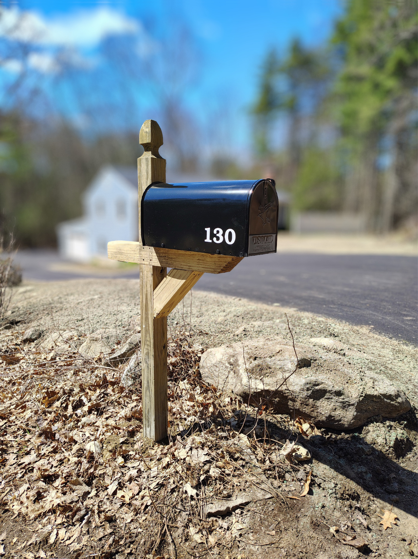 Black mailbox with the number 130, mounted on a wooden post, located on a patch of dirt and surrounded by fallen leaves, with a blurred house and trees in the background.