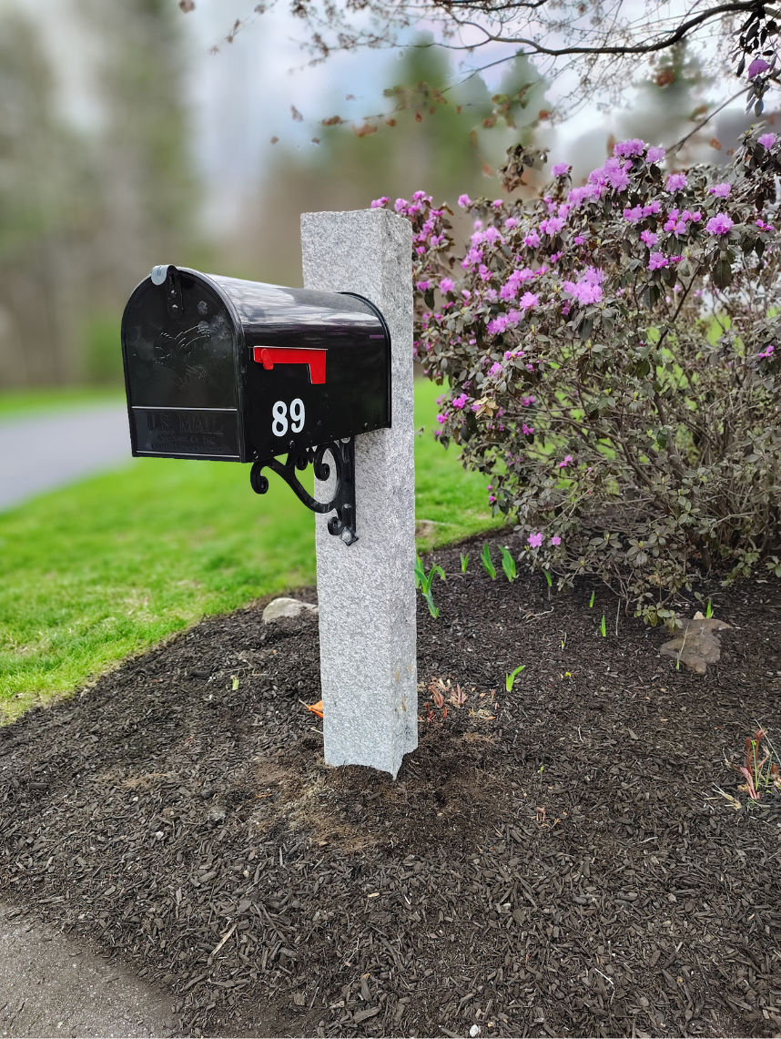 Black mailbox with red flag and number 89 on a gray stone post next to a flowering bush with purple flowers in a landscaped yard.