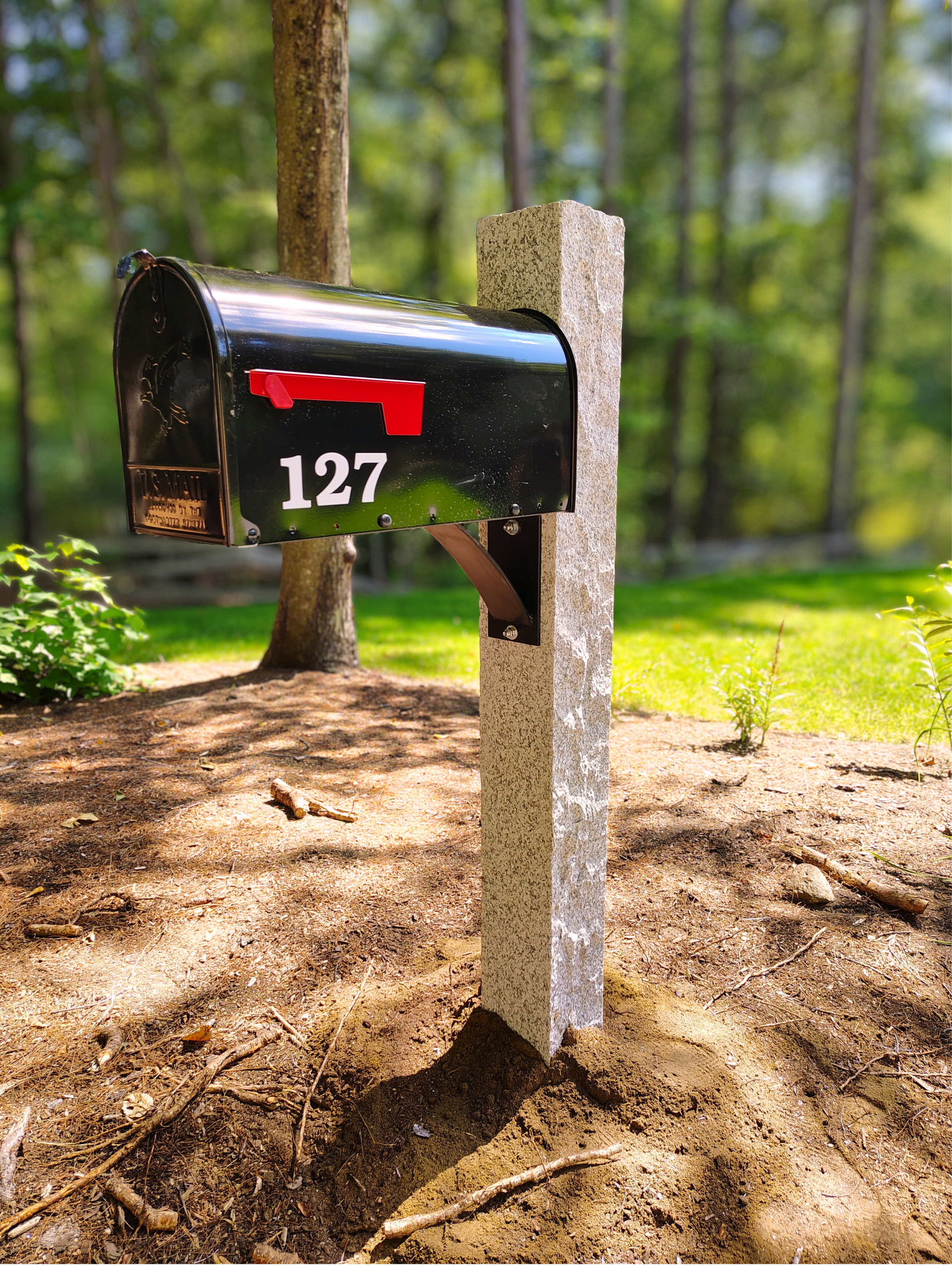 Black mailbox with red flag, labeled with the number 127, mounted on a concrete post in a wooded yard.