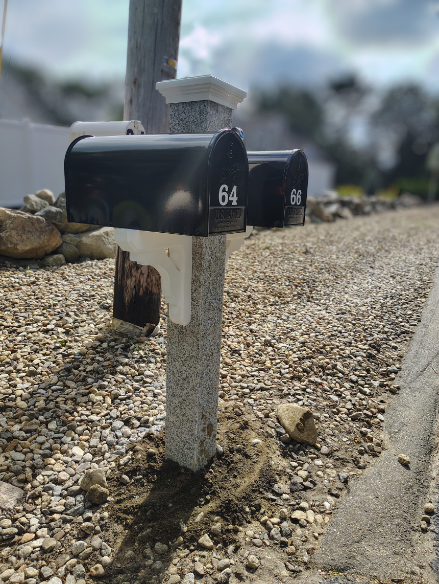 two black mailboxes mounted to a granite mailbox post by two white wood brackets. with a white cap