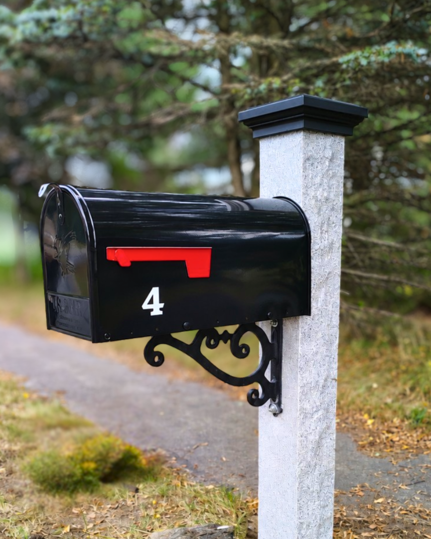 Black mailbox with a red flag mounted on a white stone post along a sidewalk in a suburban neighborhood, with trees in the background.