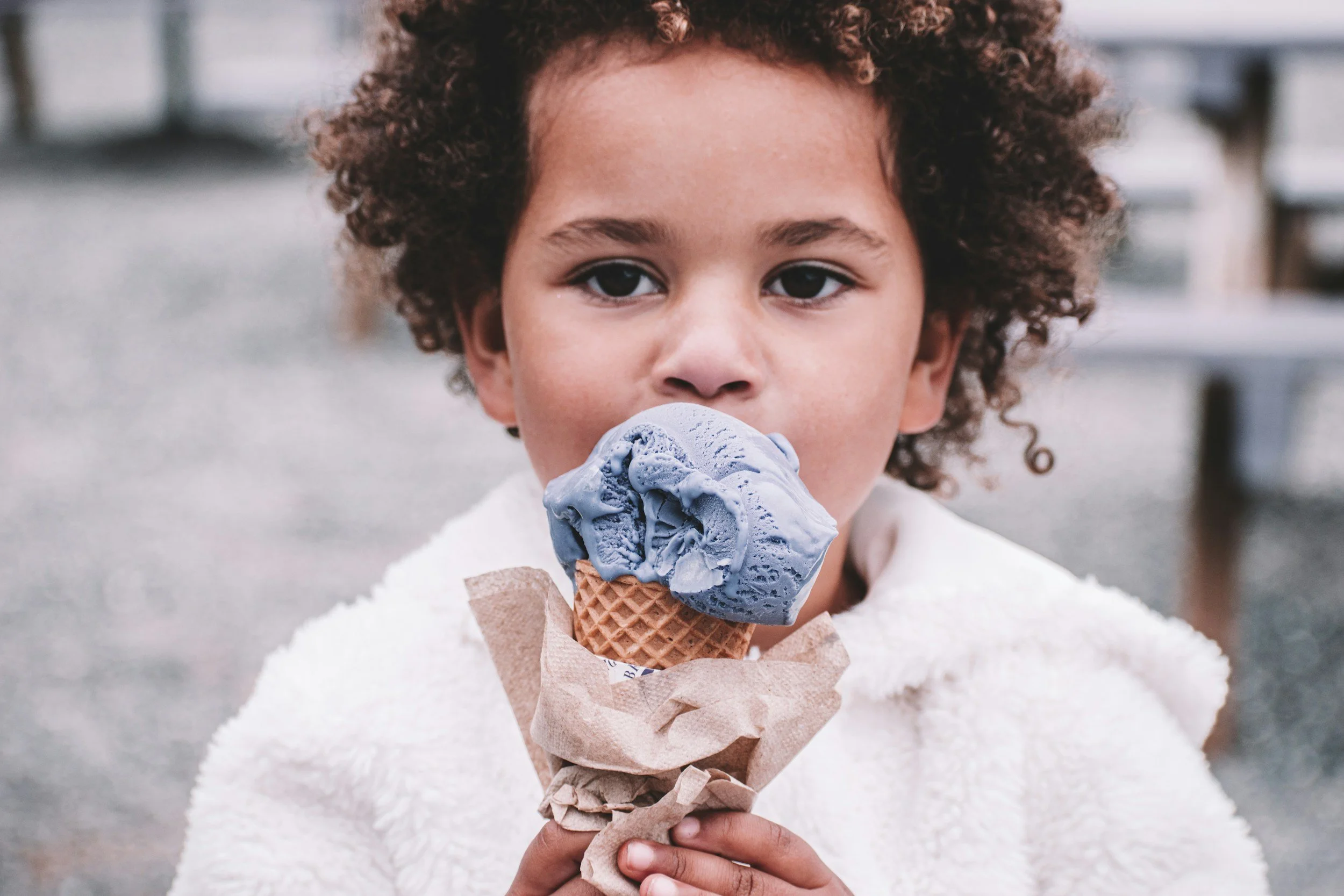 Young child with curly hair eating a blue ice cream cone outdoors.
