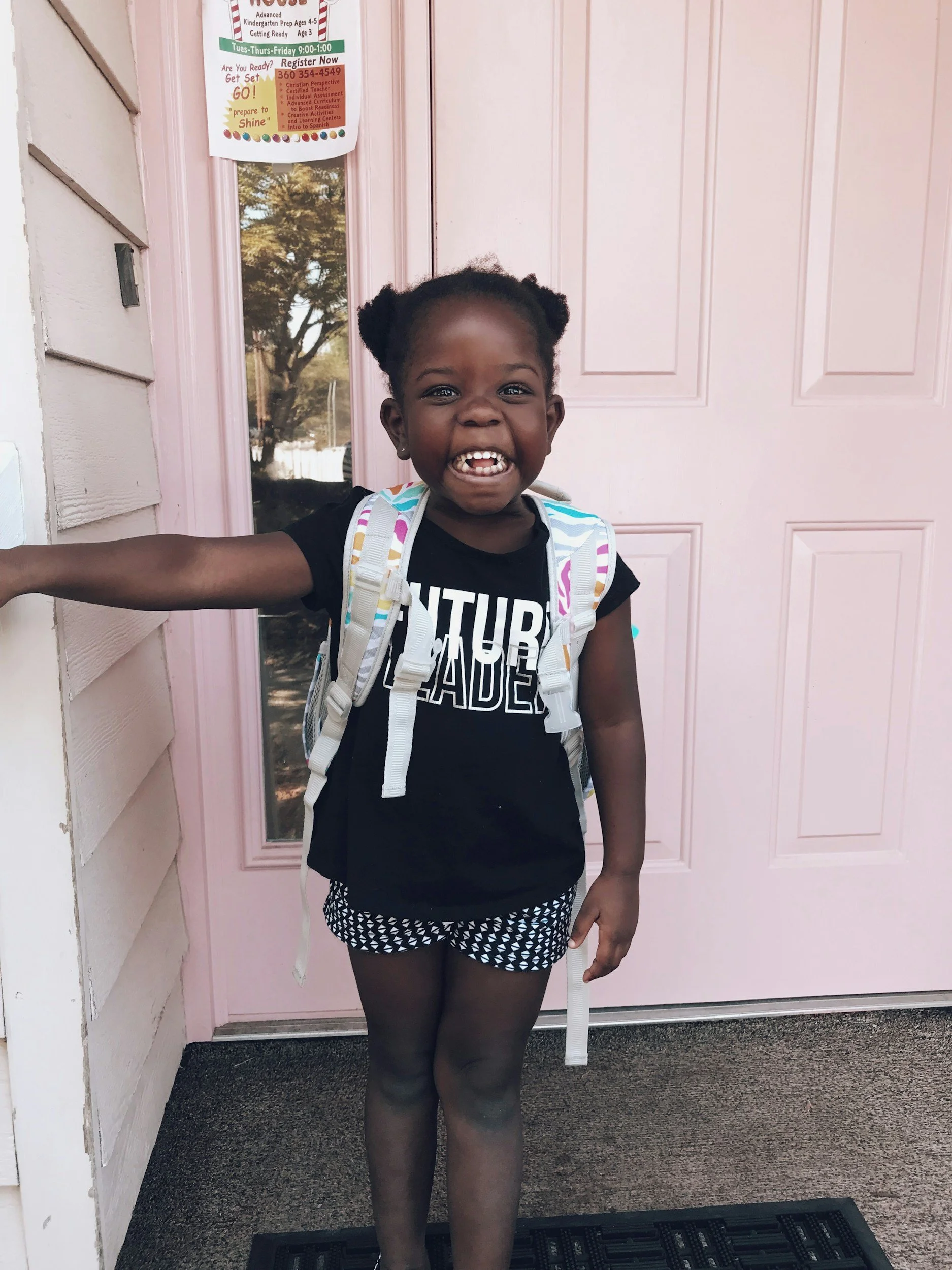A joyful young girl with pigtails is standing in front of a pink front door, smiling broadly with her teeth visible. She is wearing a black t-shirt with white text, patterned shorts, and a backpack.